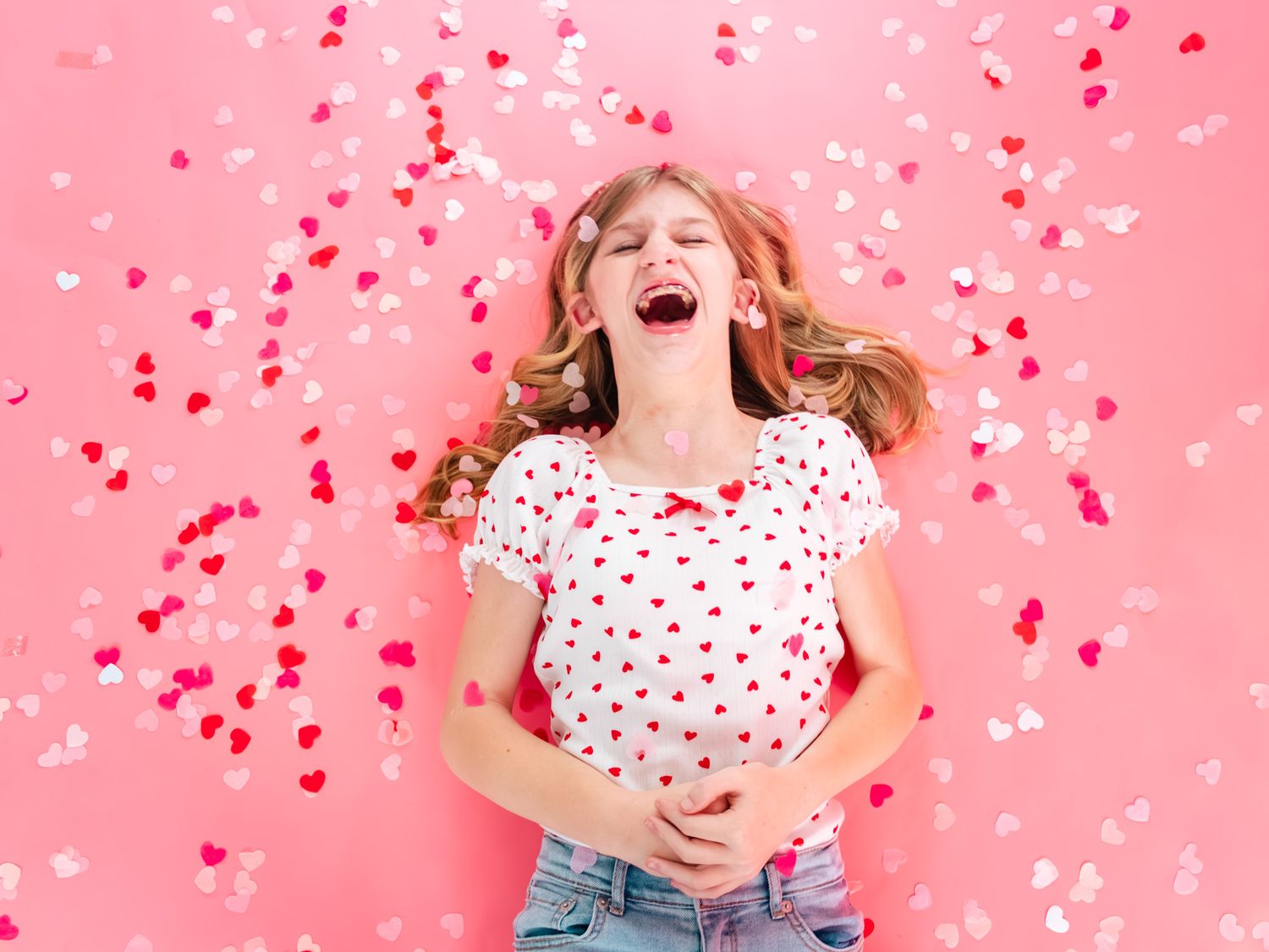 Joyful young woman laughing with heart confetti falling around her against a pink background.