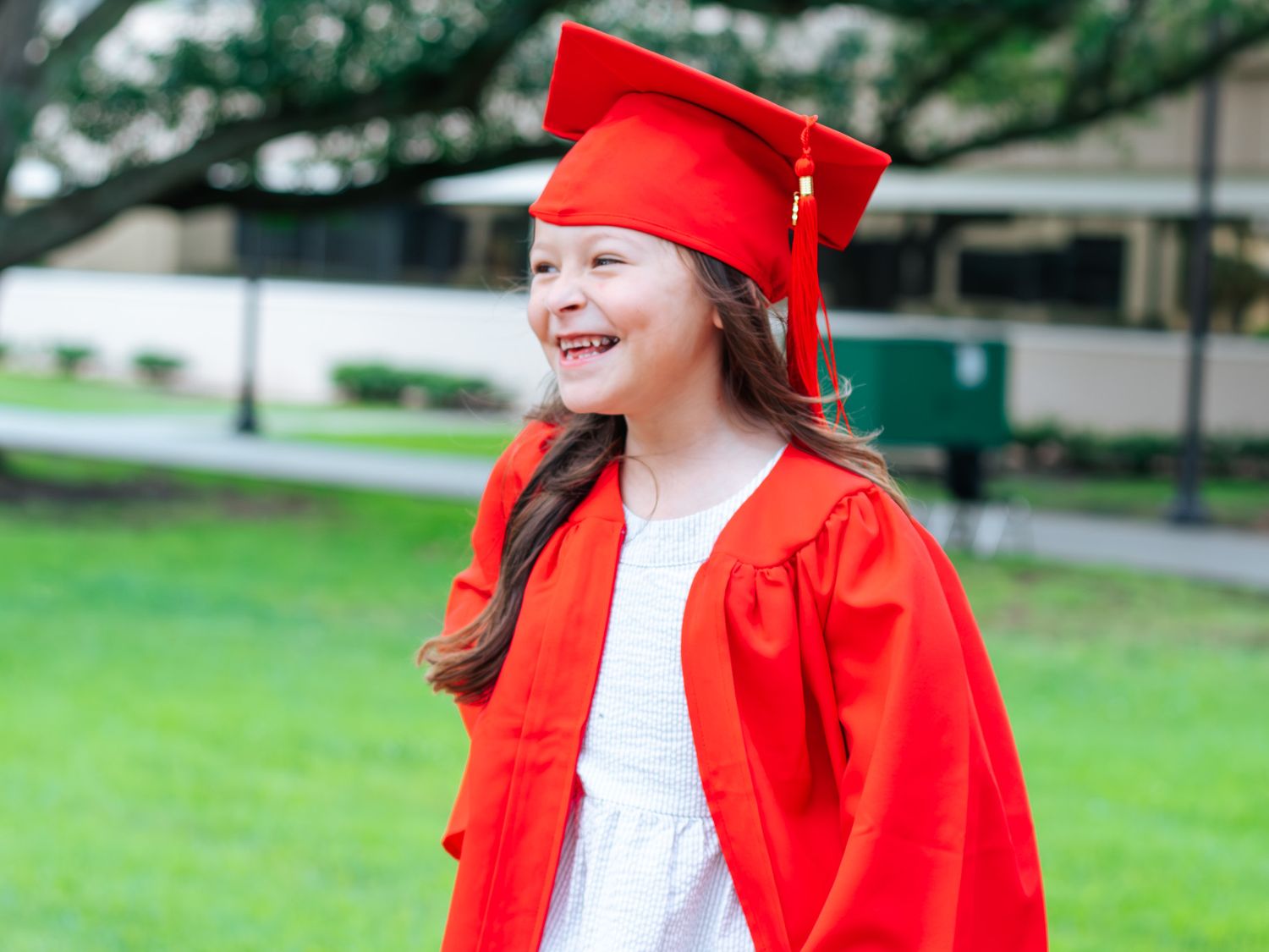 Young girl smiling in red graduation cap and gown outdoors on green grass.