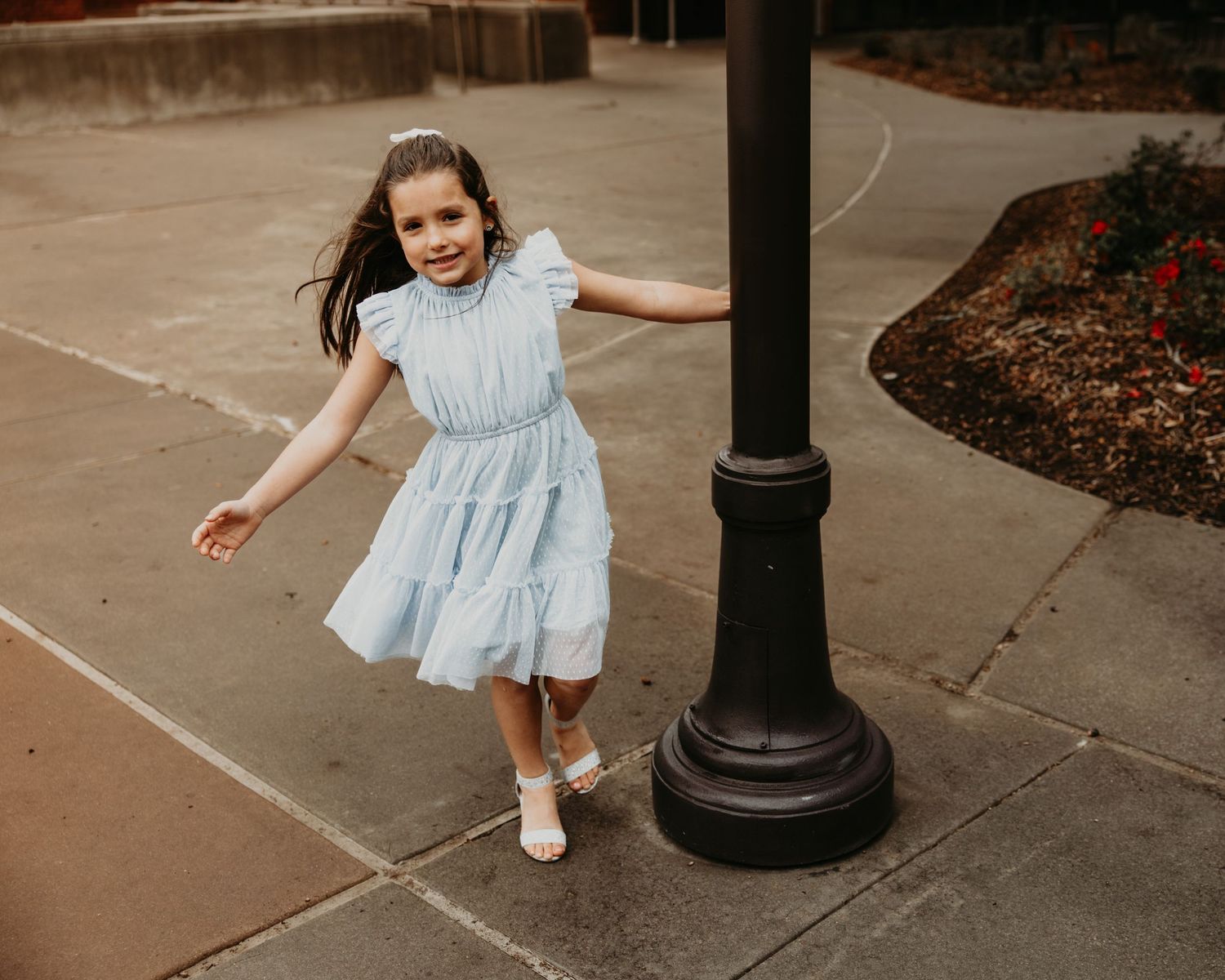 Child playing and twirling in light blue dress on concrete pathway near lamp post.