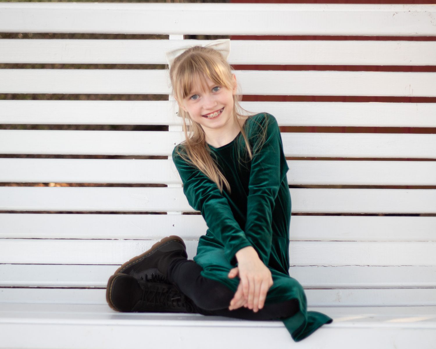Young girl in green velvet dress sitting on white bench, smiling, wearing white bow headband.
