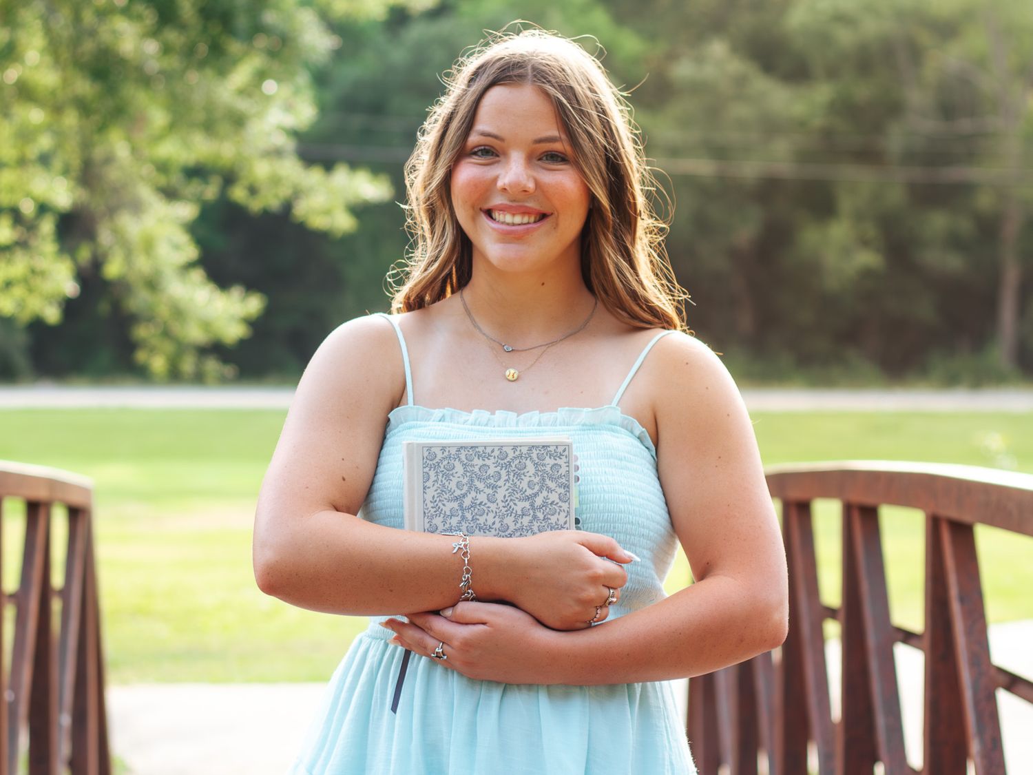 Young woman in light blue dress holding silver clutch, smiling outdoors near wooden bridge.