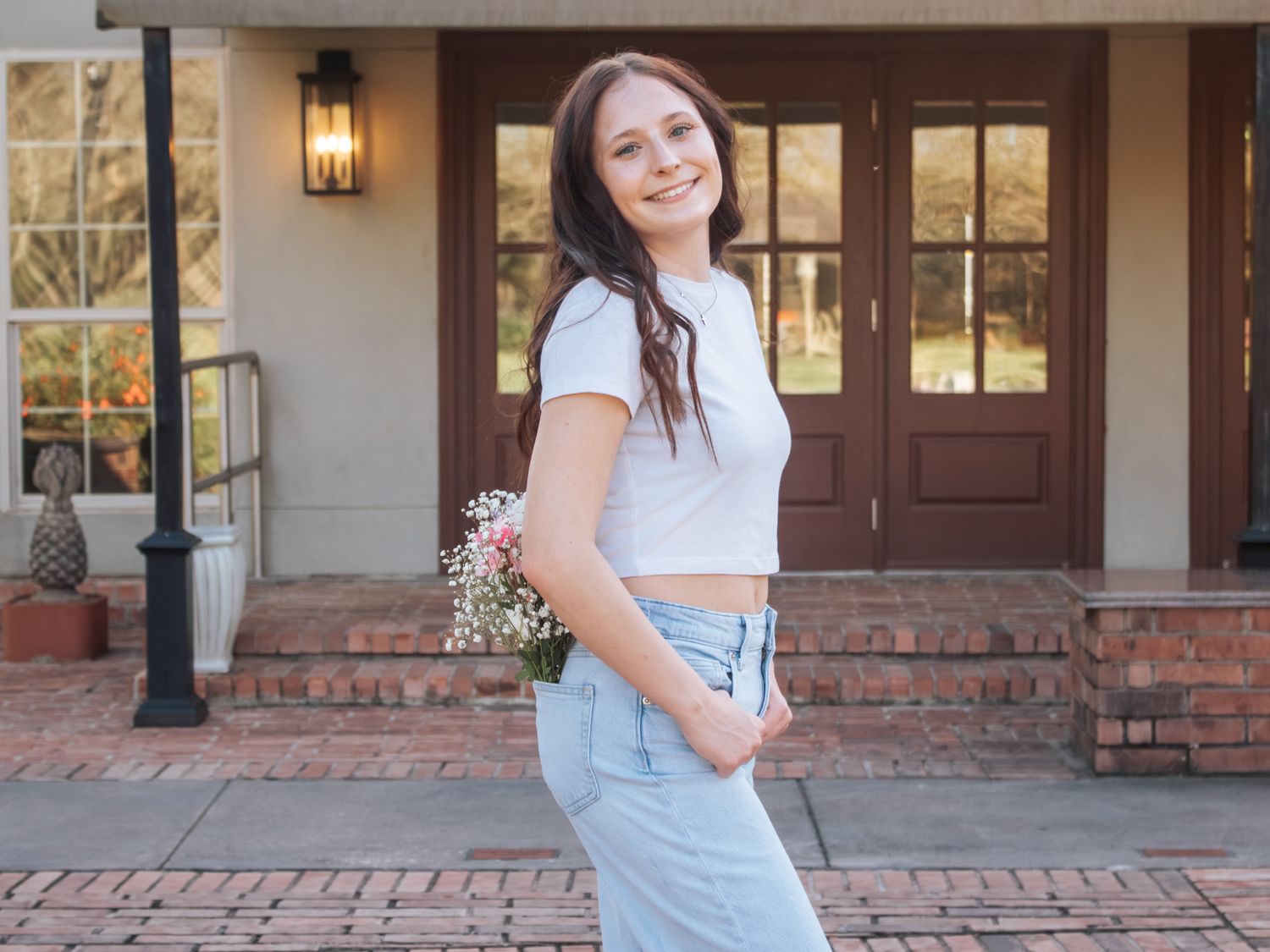 Young woman in white crop top and jeans smiling outdoors, holding wildflowers in front of a brick home.