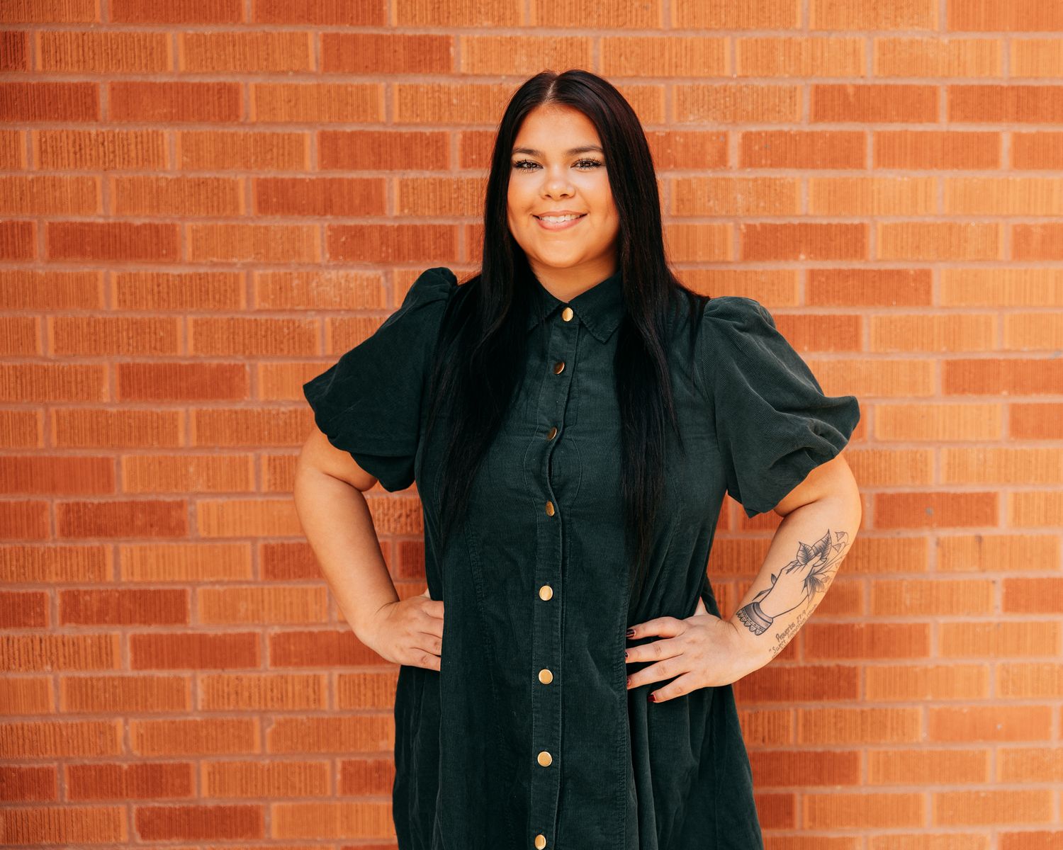 Woman in dark green button-up dress with puff sleeves, smiling confidently against a brick wall backdrop.