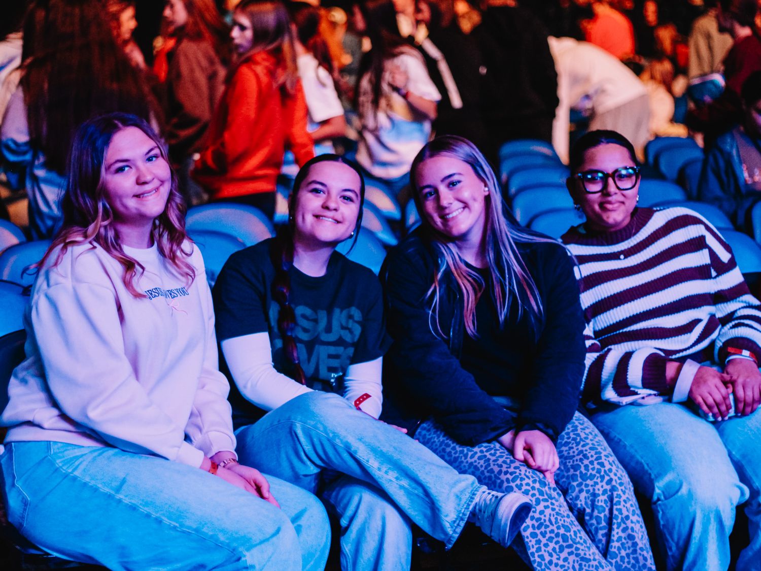 Four young women smiling and sitting together in blue stadium seats at a live event.