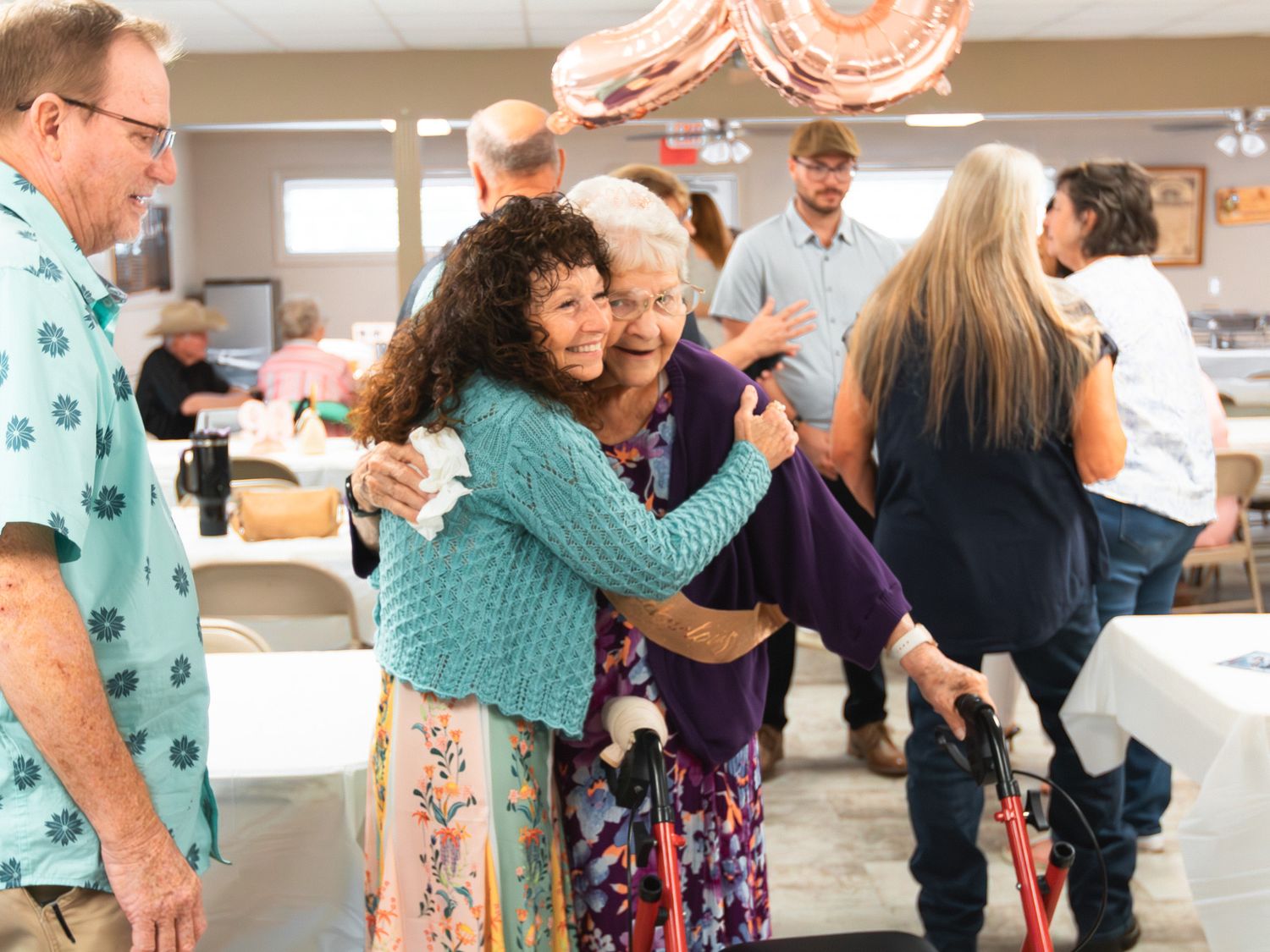 Two women share a warm hug at a festive indoor gathering with pink balloons and guests in the background.