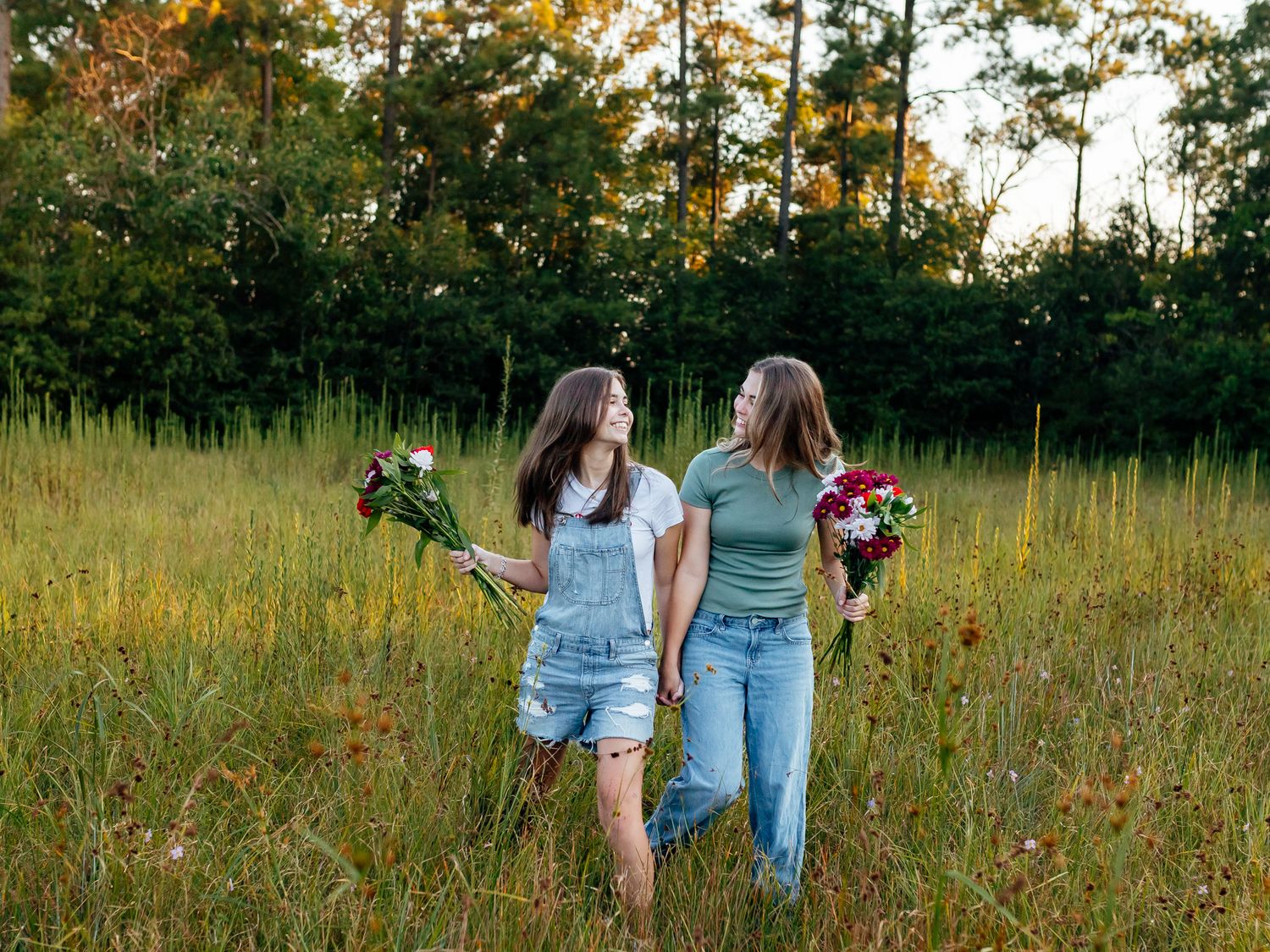 Two young women holding bouquets walk hand in hand through a lush green meadow at golden hour.