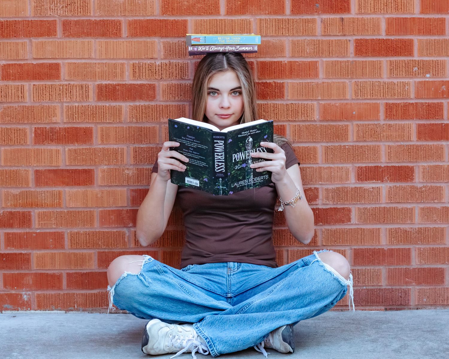 Person in jeans sits cross-legged against brick wall reading a dark-colored book in multiple poses.