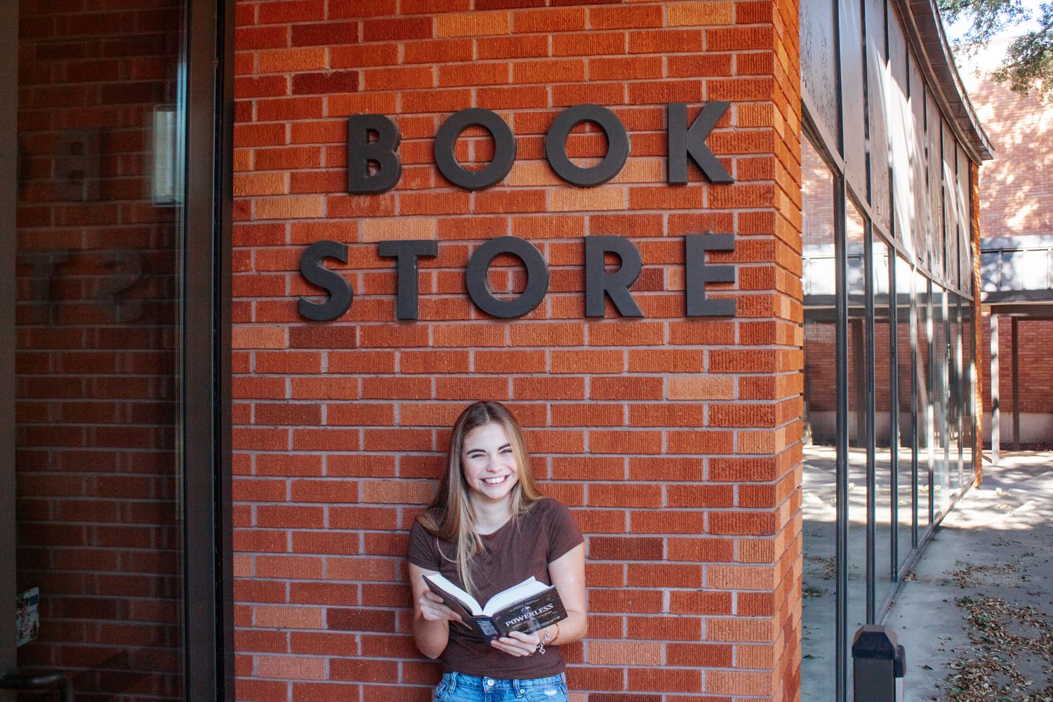 Smiling young woman holding a book outside a brick bookstore building with large black letter signage.