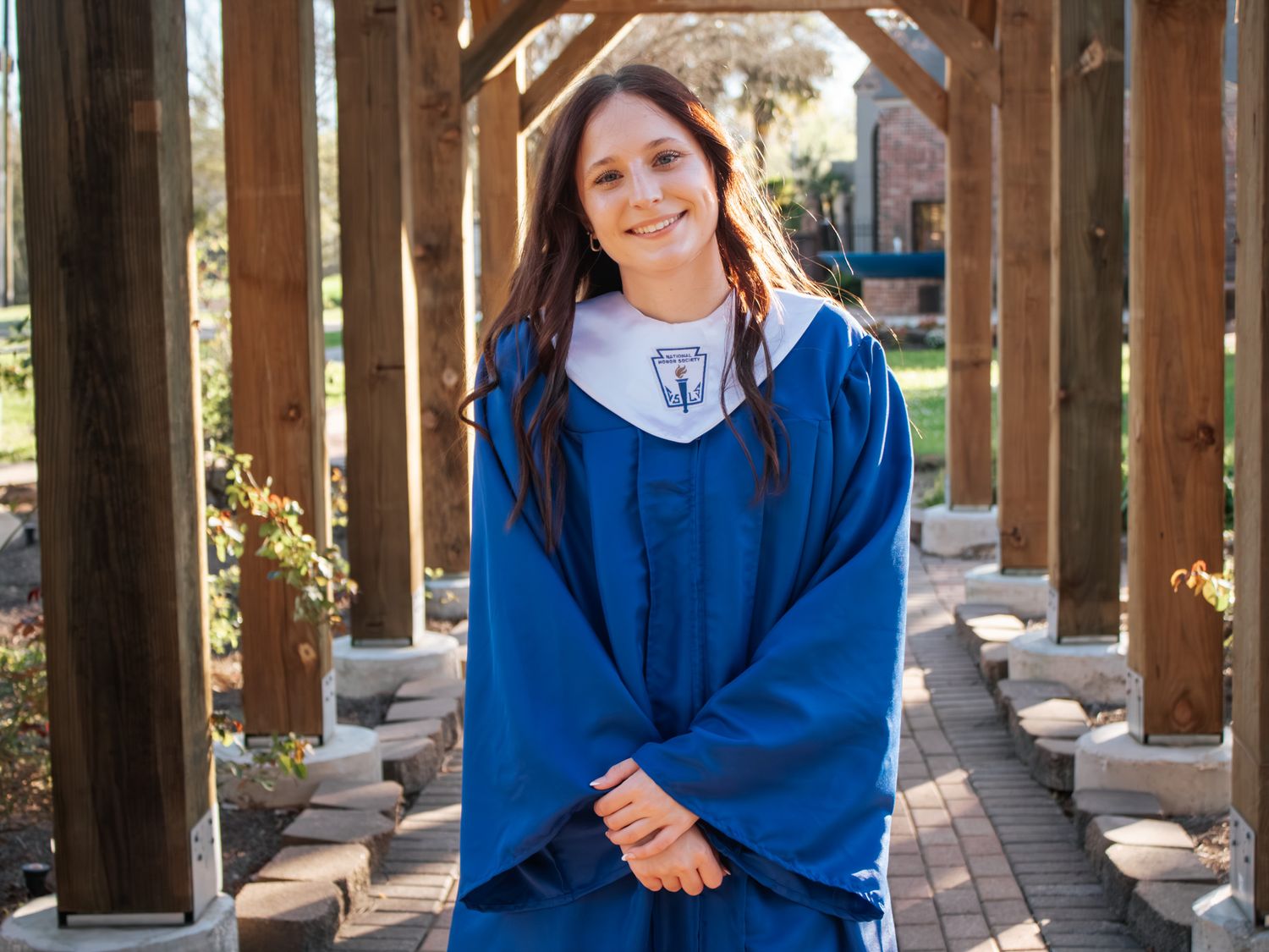 Smiling graduate in blue cap and gown posing under wooden pergola on sunny day.