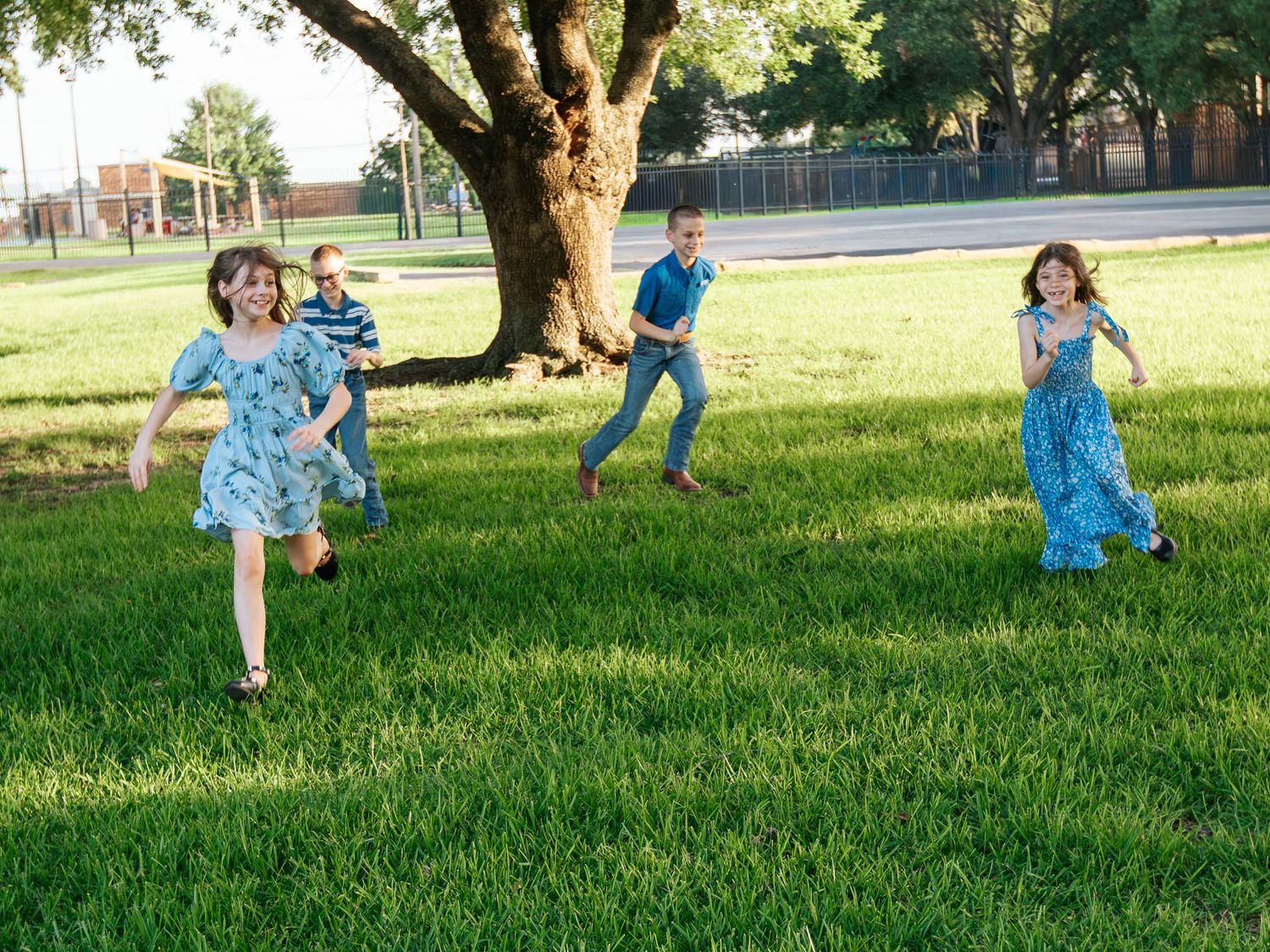 Four children running and playing on lush green grass near a large tree on a sunny day.