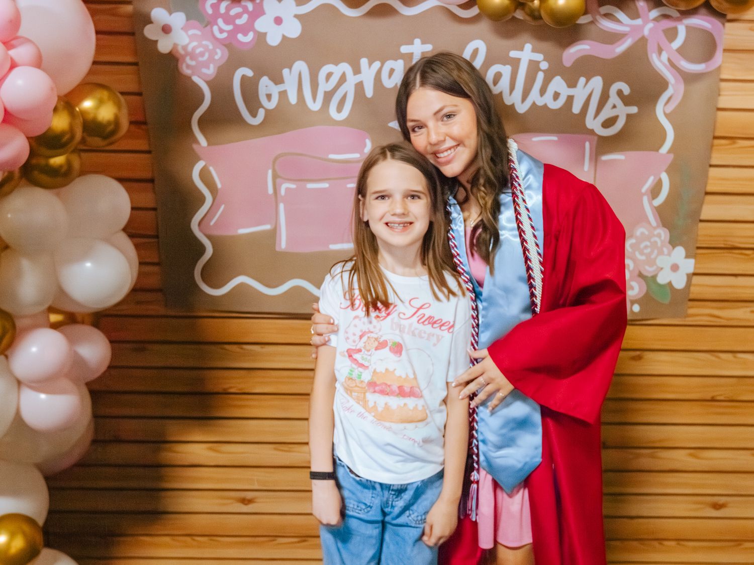 A young girl poses with a graduating woman in red cap and gown in front of a Congratulations balloon backdrop.