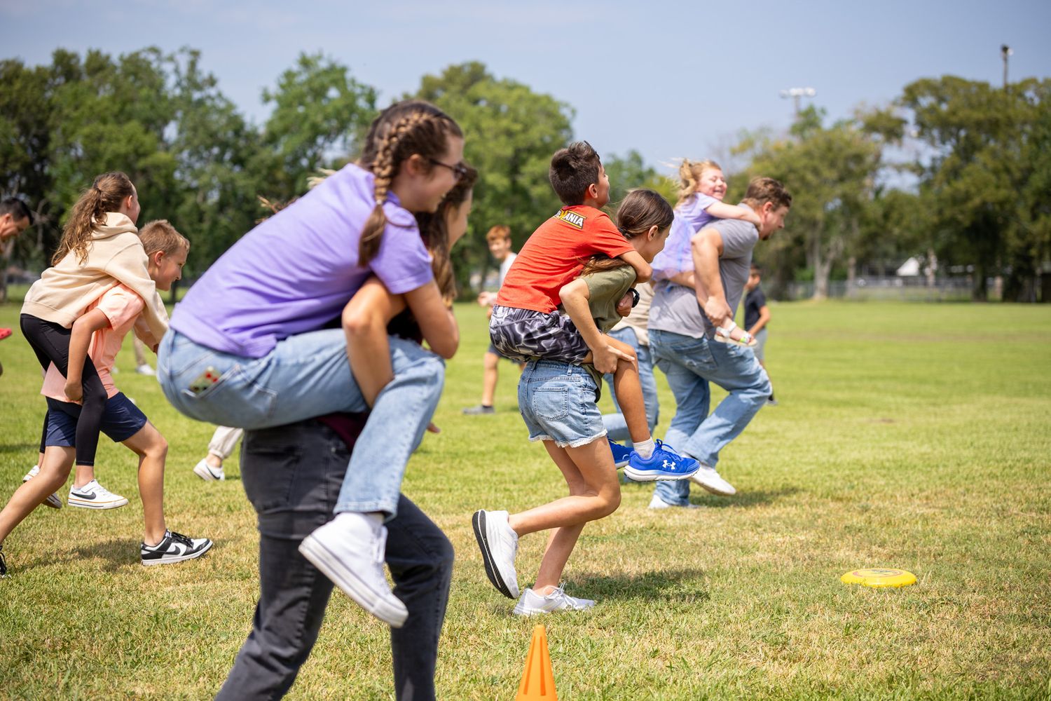 Children playing piggyback race game on green grass field during sunny outdoor school activity day.
