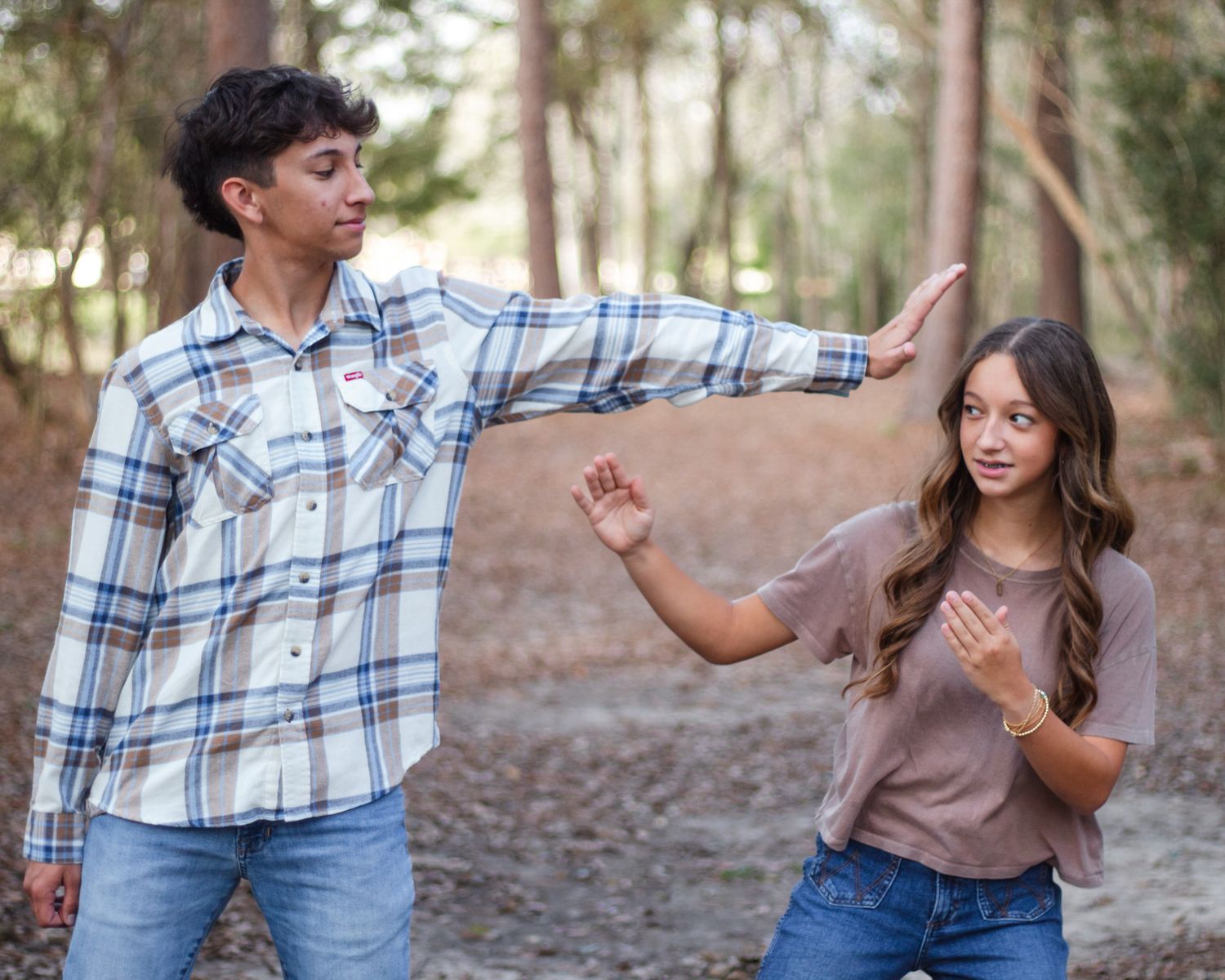 Two teens playfully dancing outdoors among pine trees, boy in plaid shirt gesturing toward girl in mauve top and jeans.