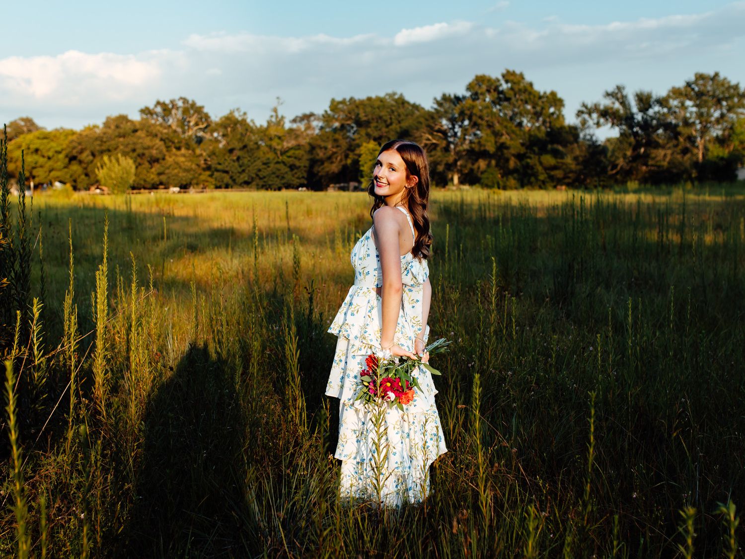 Bride in white tiered lace dress holding red bouquet standing in lush green meadow at golden hour.