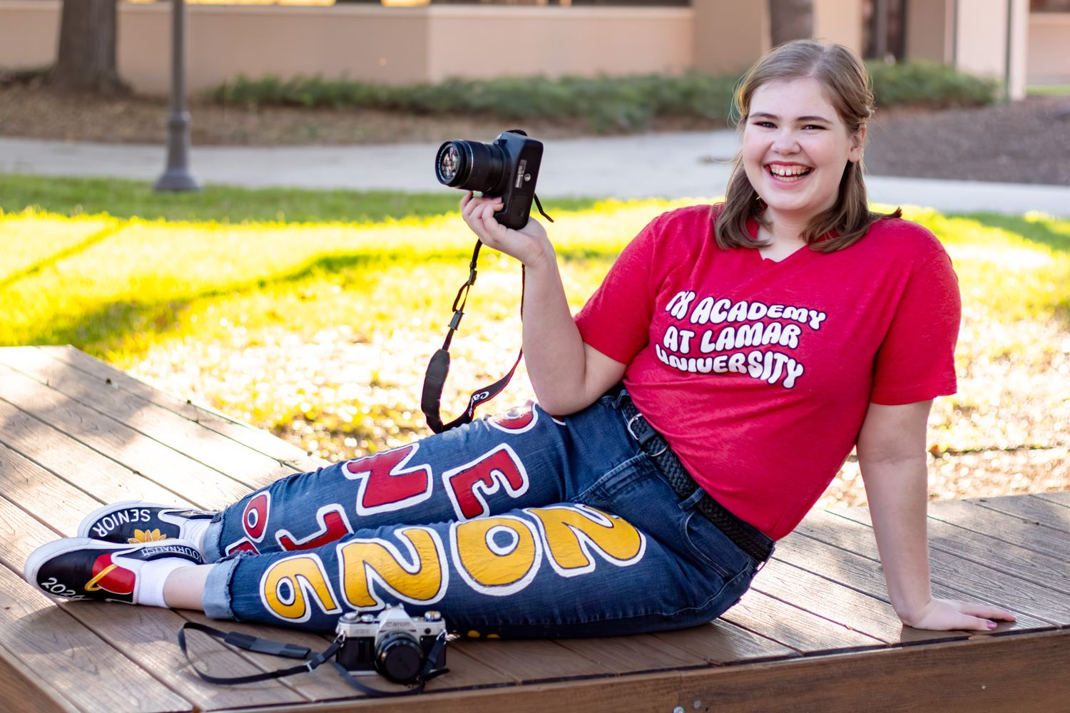 Smiling woman in red Academy at Lamar University t-shirt holding camera, sitting on bench in jeans.