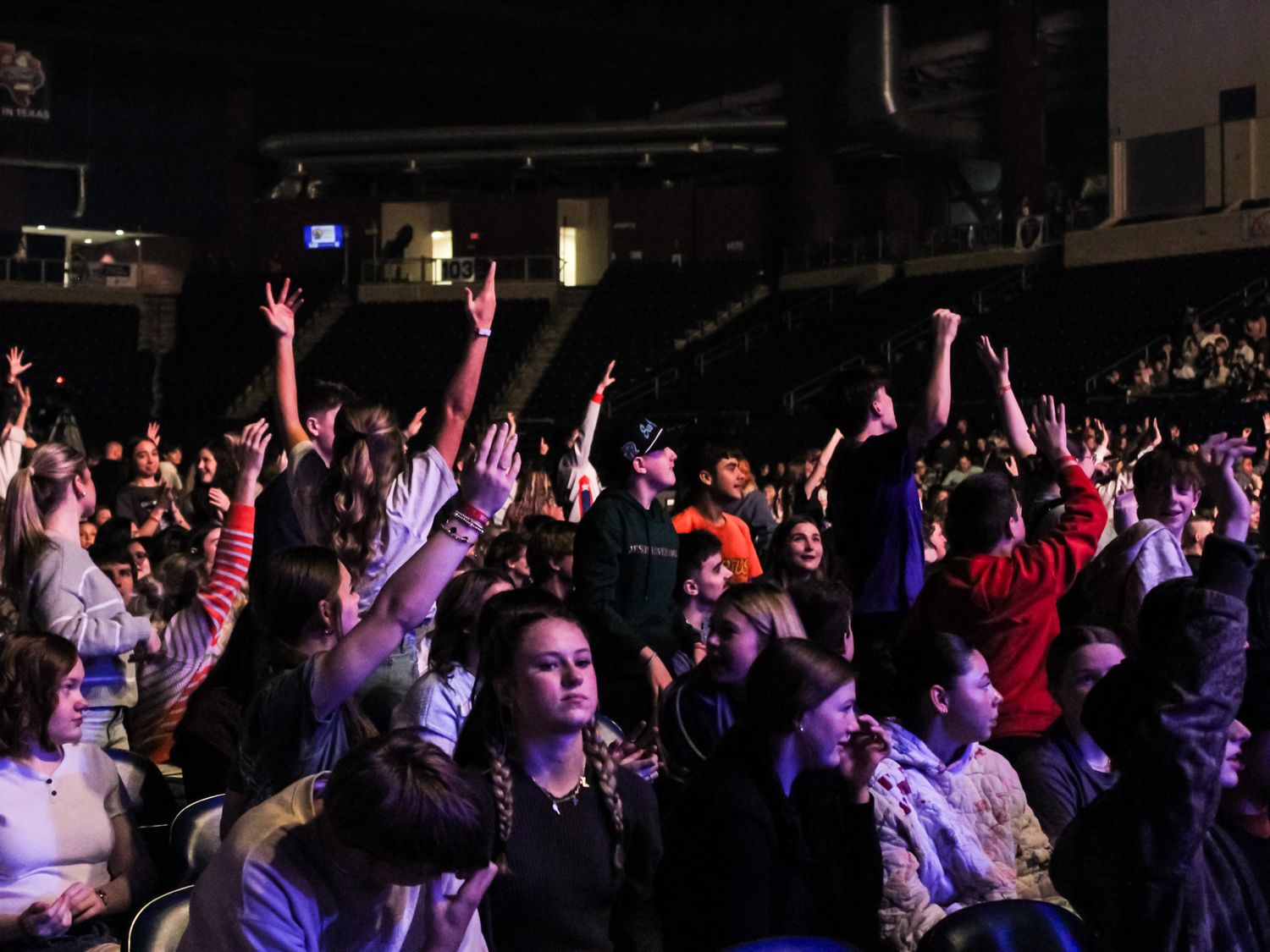 Excited crowd of young people with hands raised at a live indoor concert or performance event.