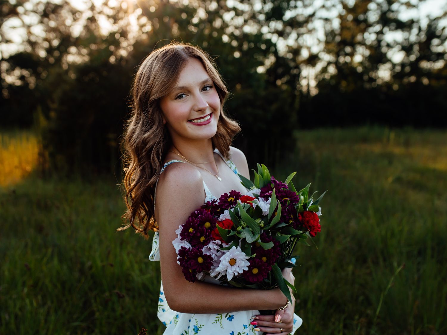 Young woman smiling outdoors at golden hour, holding a bouquet of red and white flowers in a floral dress.