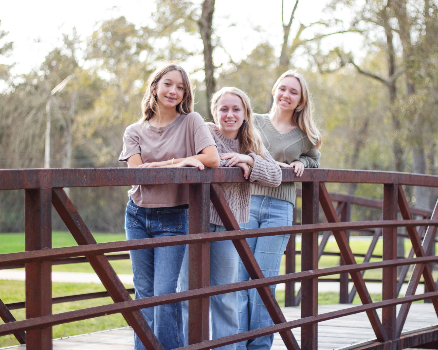 Three teenage girls smiling on a wooden bridge outdoors, wearing casual tops and jeans, surrounded by trees.