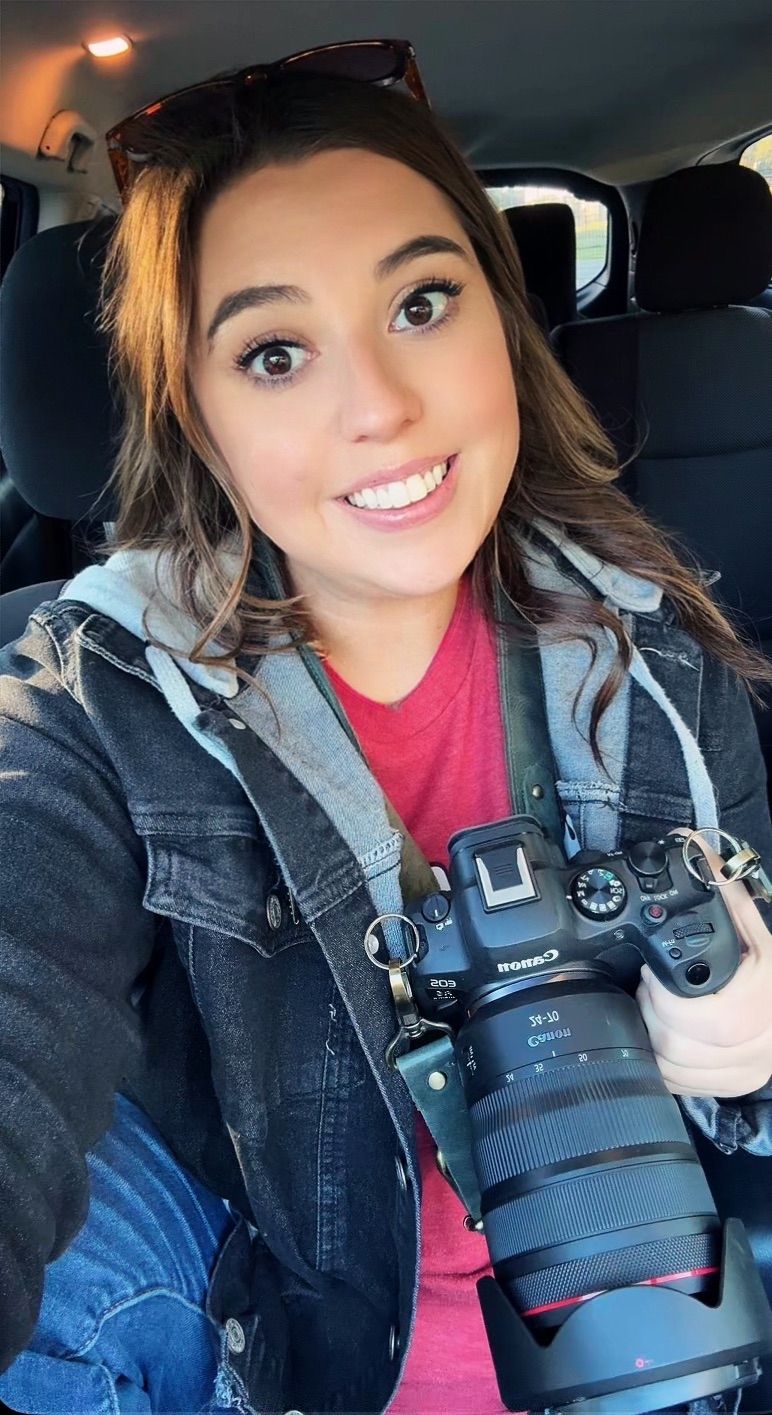 A photographer wearing a denim jacket shows off a professional camera with telephoto lens while sitting in a car.