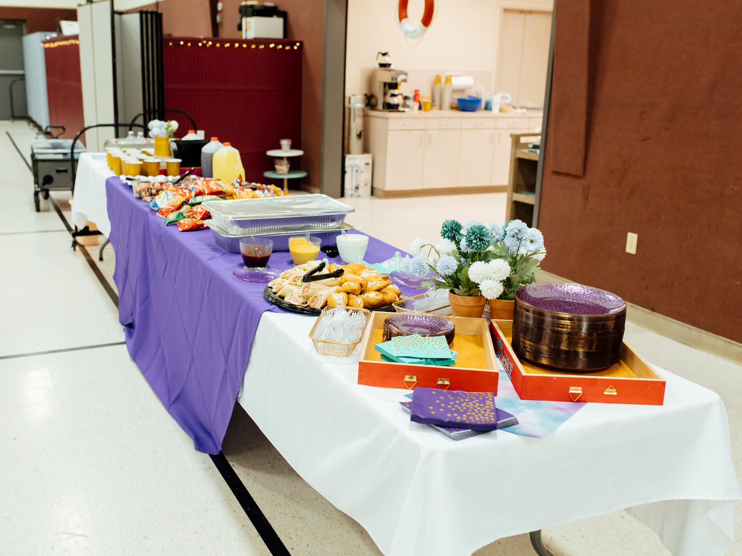 Buffet table with purple tablecloth displaying assorted food, floral centerpiece, and decorative items at an indoor event.
