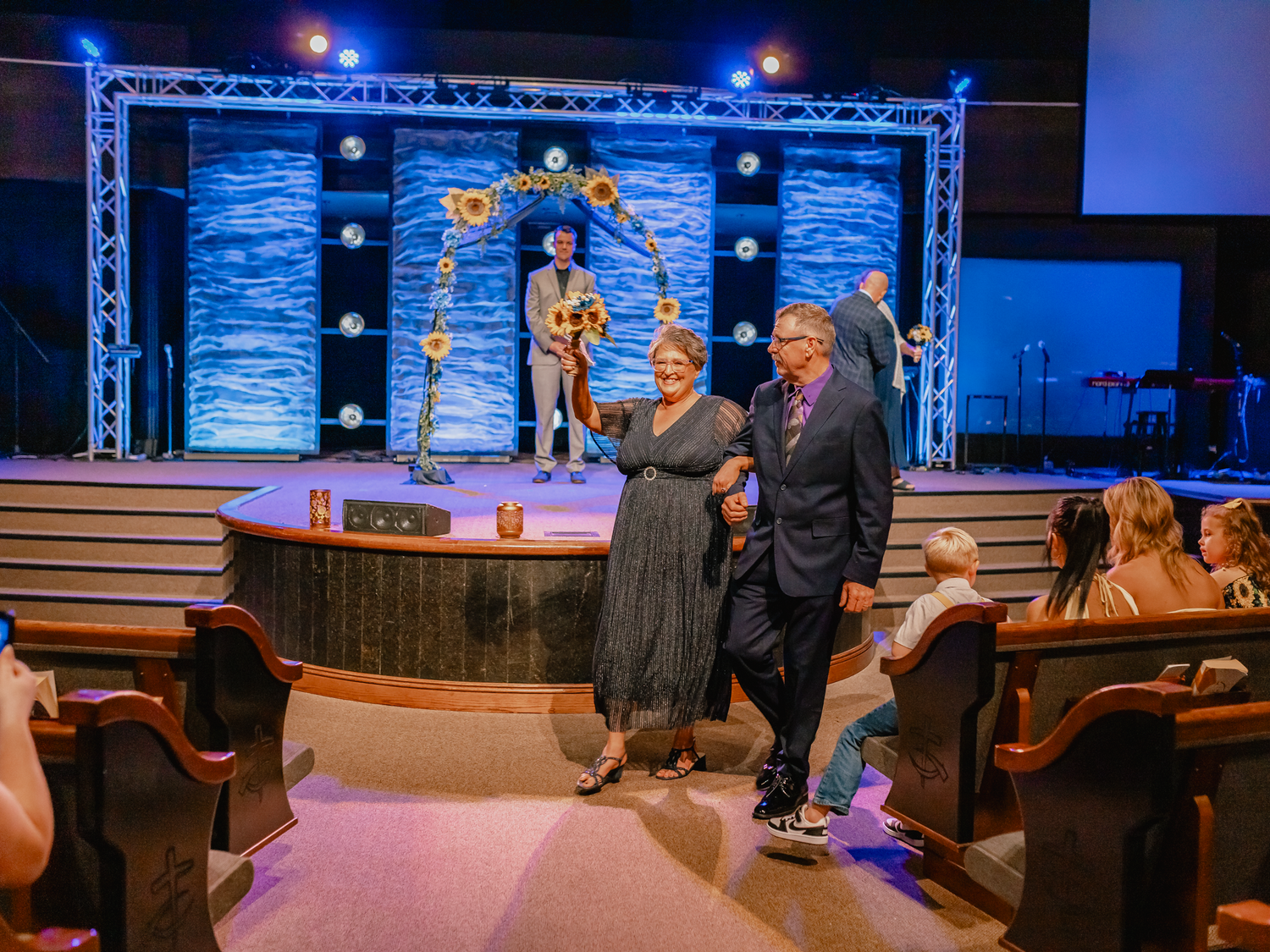 Couple dancing in church aisle during live mariachi performance on decorated stage with blue backdrop.
