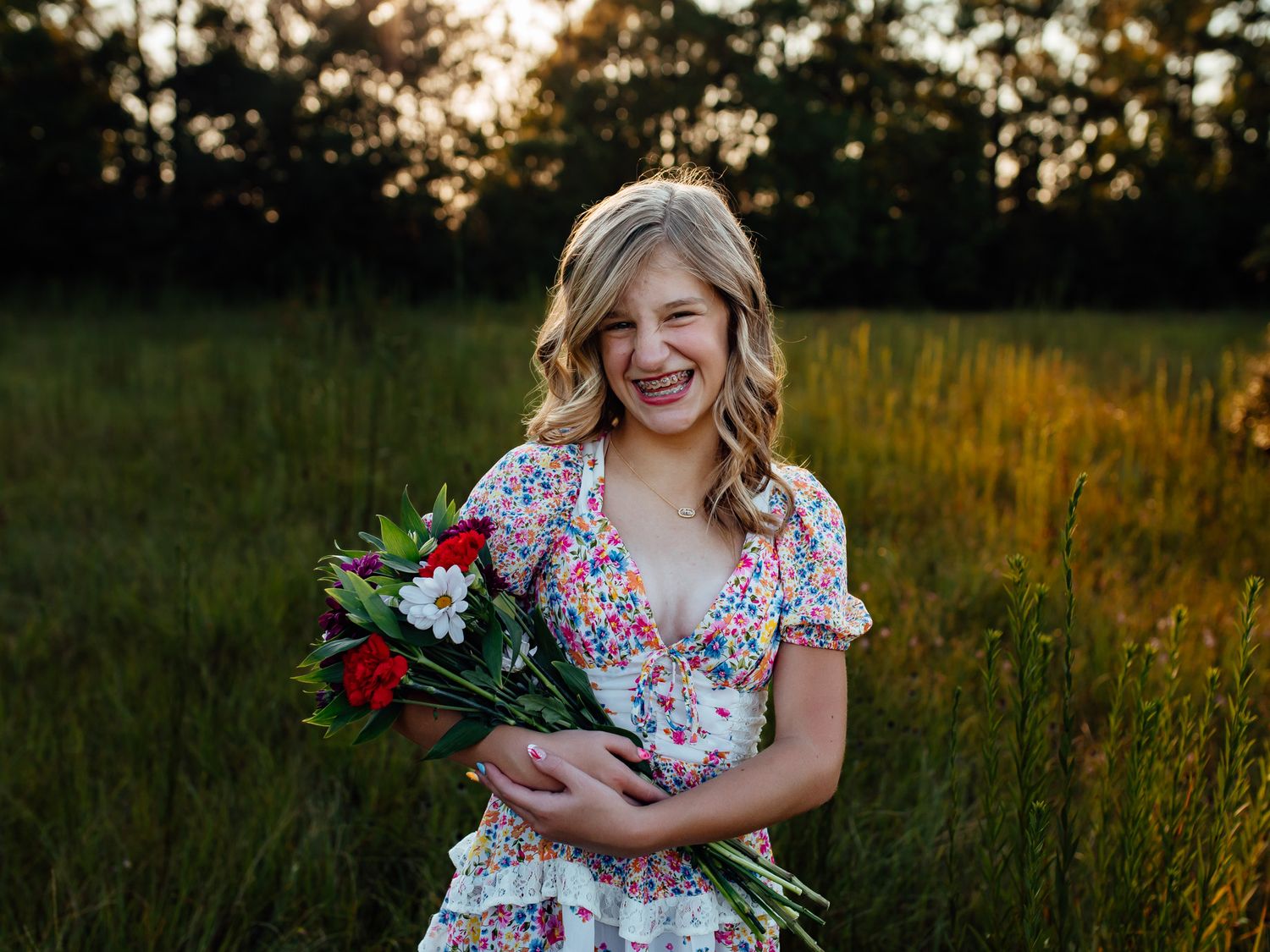 Smiling girl in floral dress holding red and white flower bouquet in sunny meadow at golden hour.