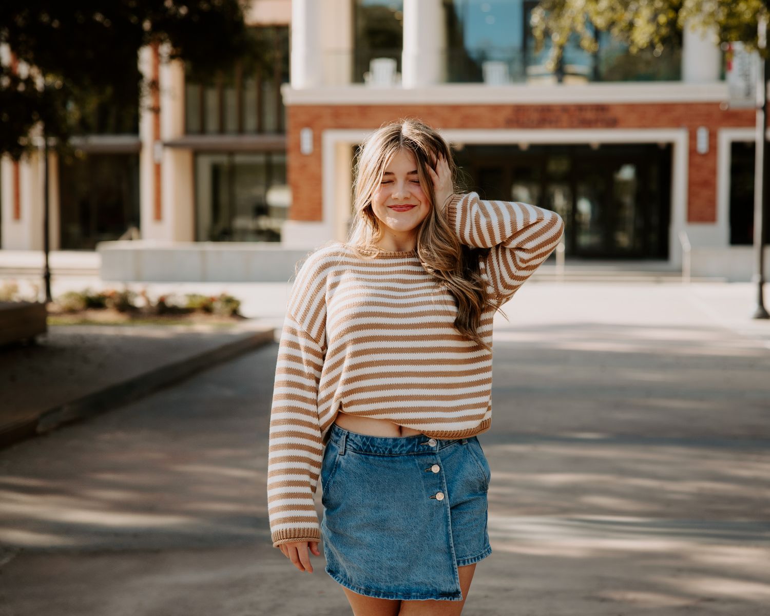 Young woman in striped sweater and denim skirt smiling outdoors near a brick building on a sunny day.
