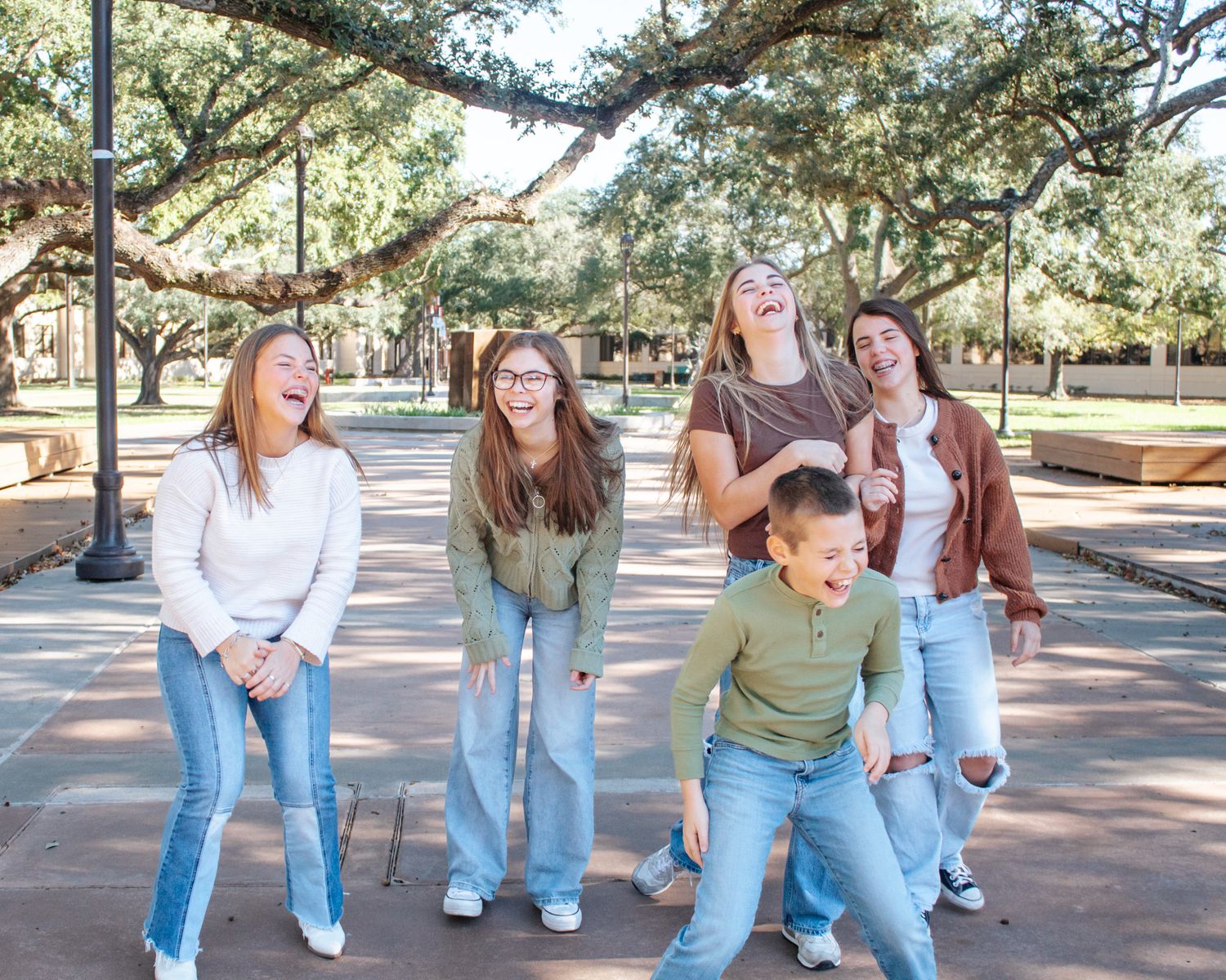 Five laughing teens in jeans enjoy a fun outdoor moment on a tree-lined campus walkway on a sunny day.