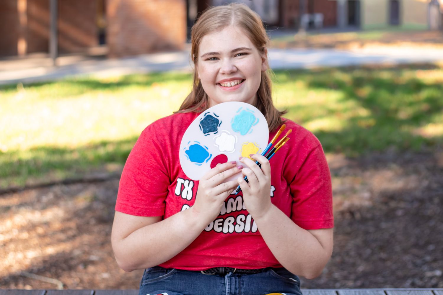 Smiling young woman in red university t-shirt holding a colorful painted soccer ball and art supplies outdoors.