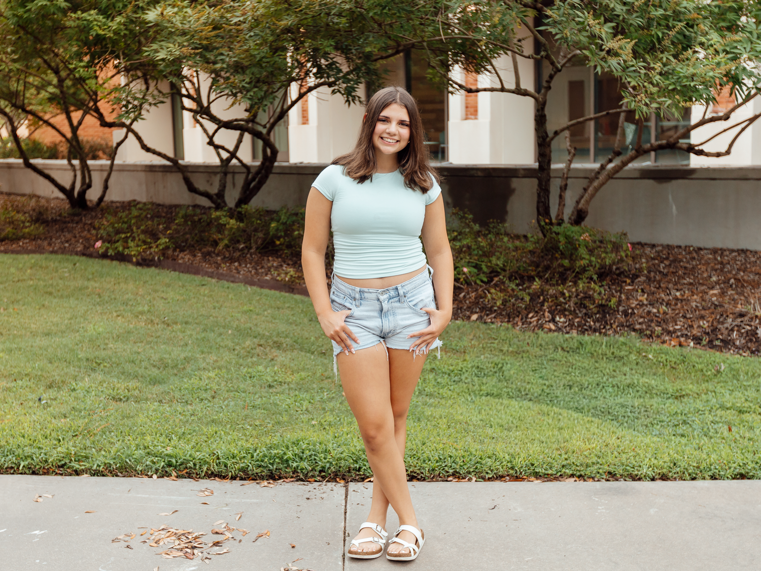 Young woman in mint crop top and denim shorts smiling outdoors on a college campus on a sunny day.