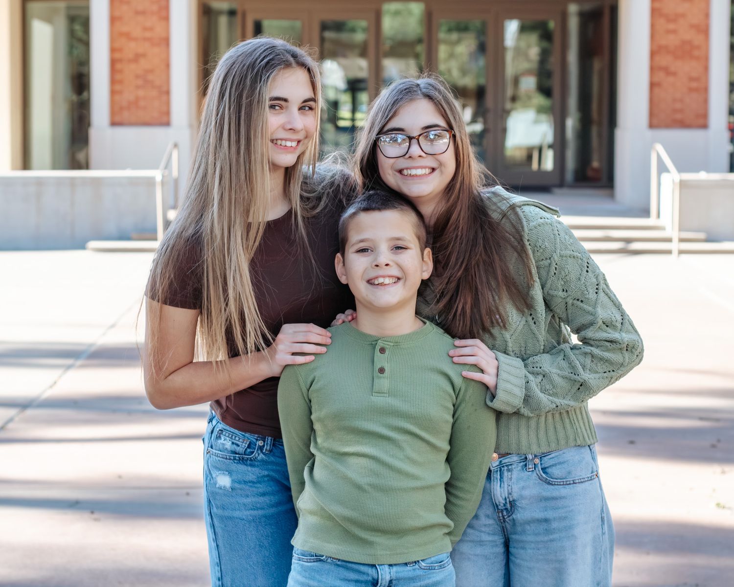 Three friends in casual clothing laughing and hugging together outdoors on a sunny day.