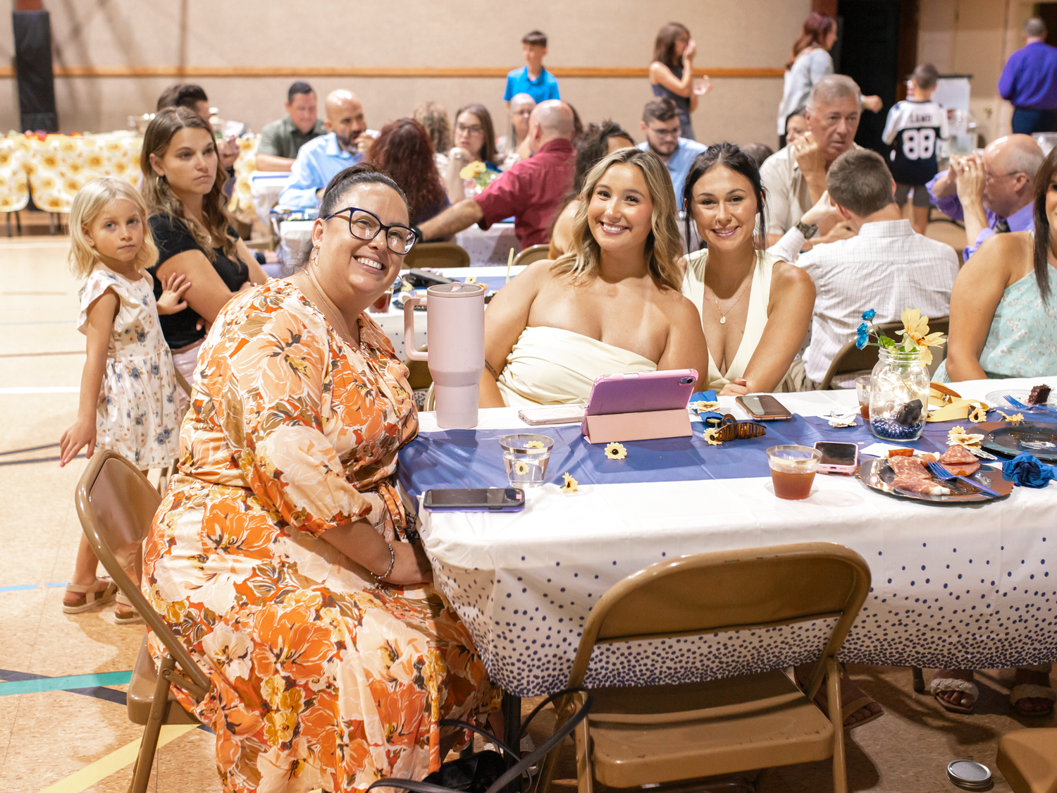Women smiling at a decorated banquet table during an indoor event in a gymnasium.