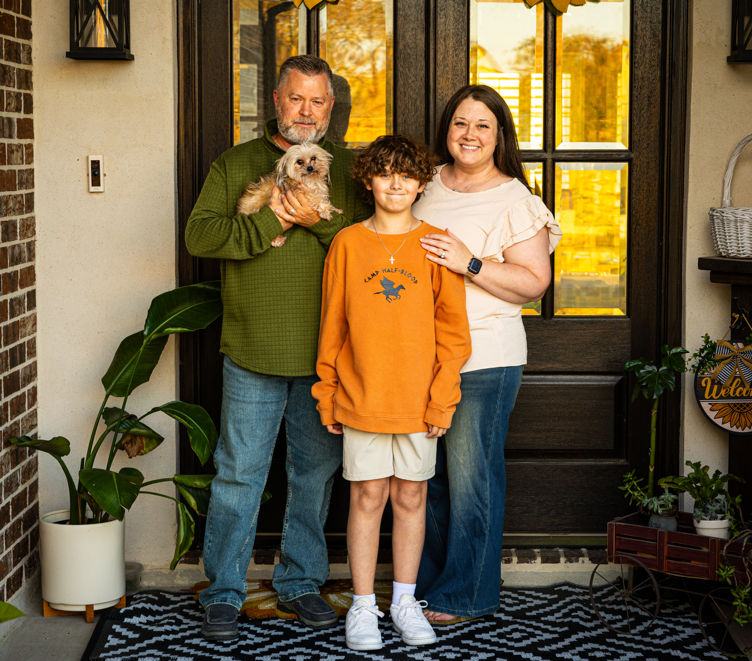A family portrait on a front porch with decorative plants and a welcome mat.