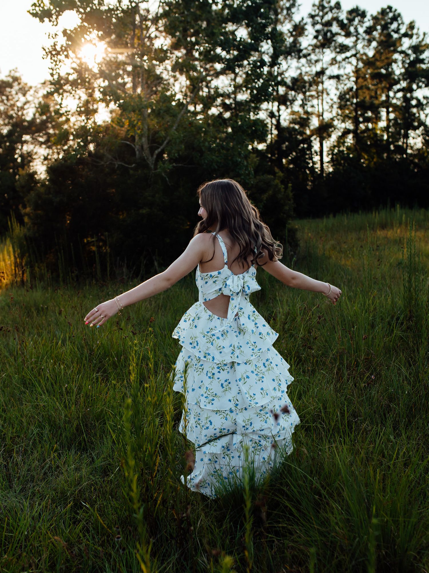 A young woman in a floral ruffled dress twirls in a sunlit green meadow at golden hour.