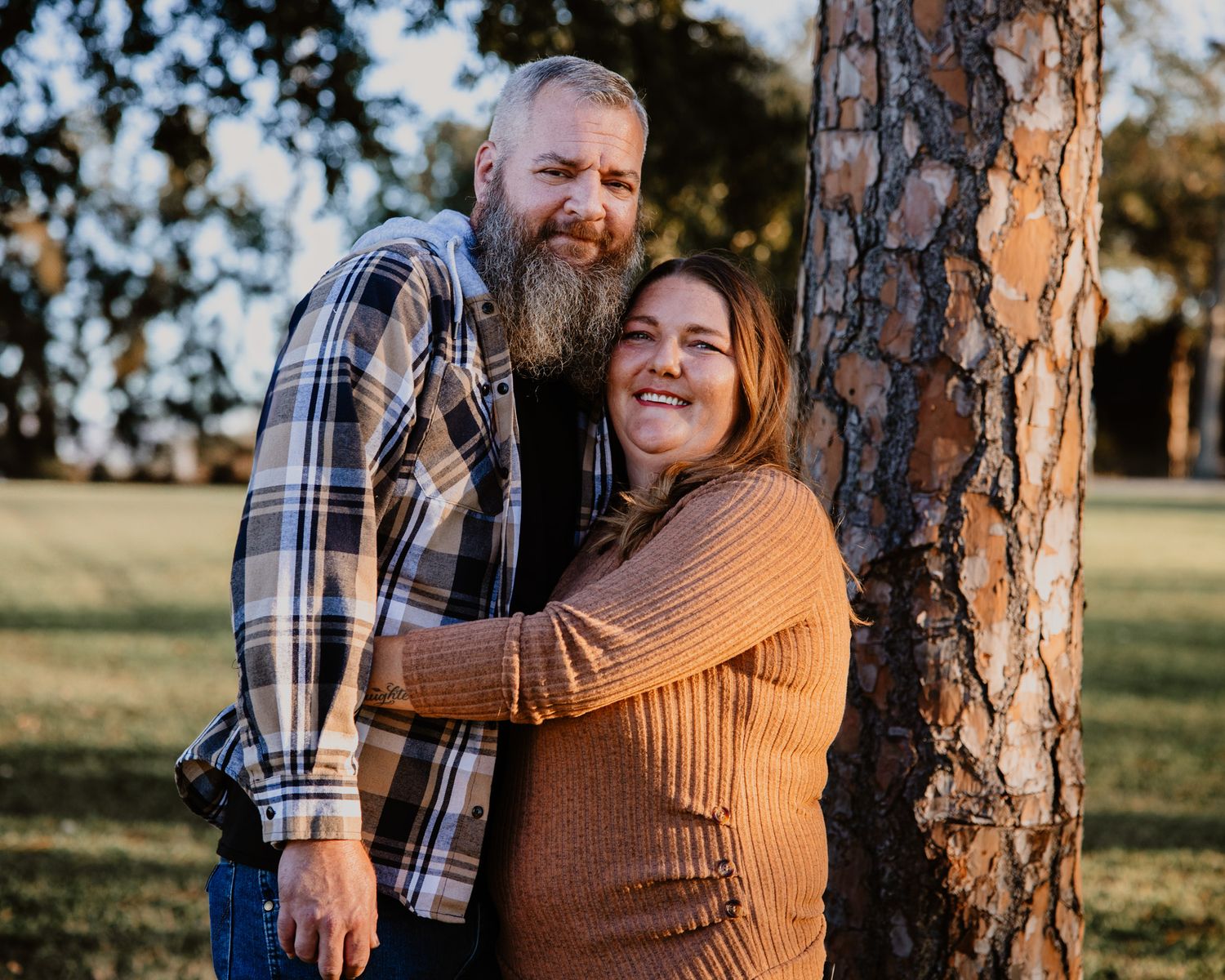 A smiling couple embraces by a tree in a park, man with beard in plaid shirt, woman in orange sweater.
