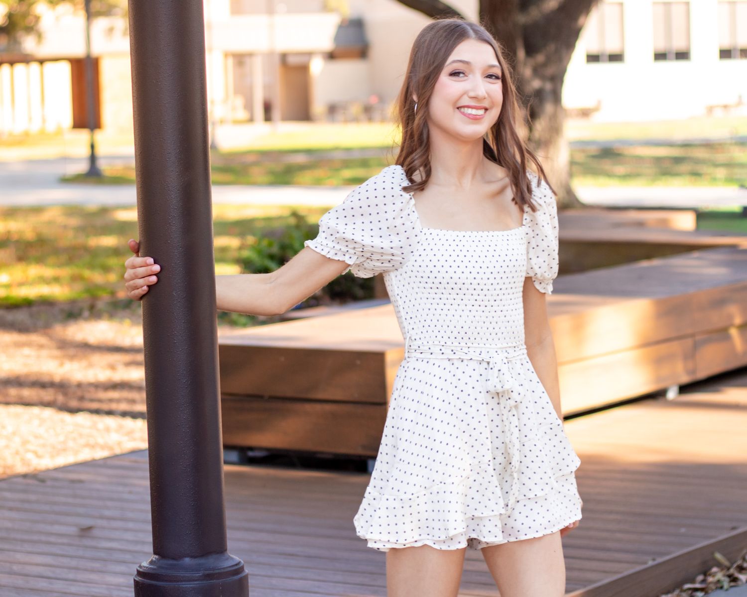 A person in a white ruffled top and shorts poses on a wooden deck with sunlit buildings in the background.