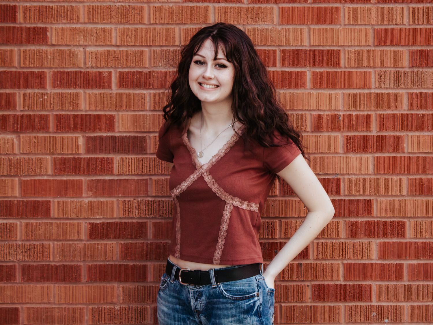 Young woman with dark curly hair smiling against a red brick wall, wearing a red V-neck top and jeans.