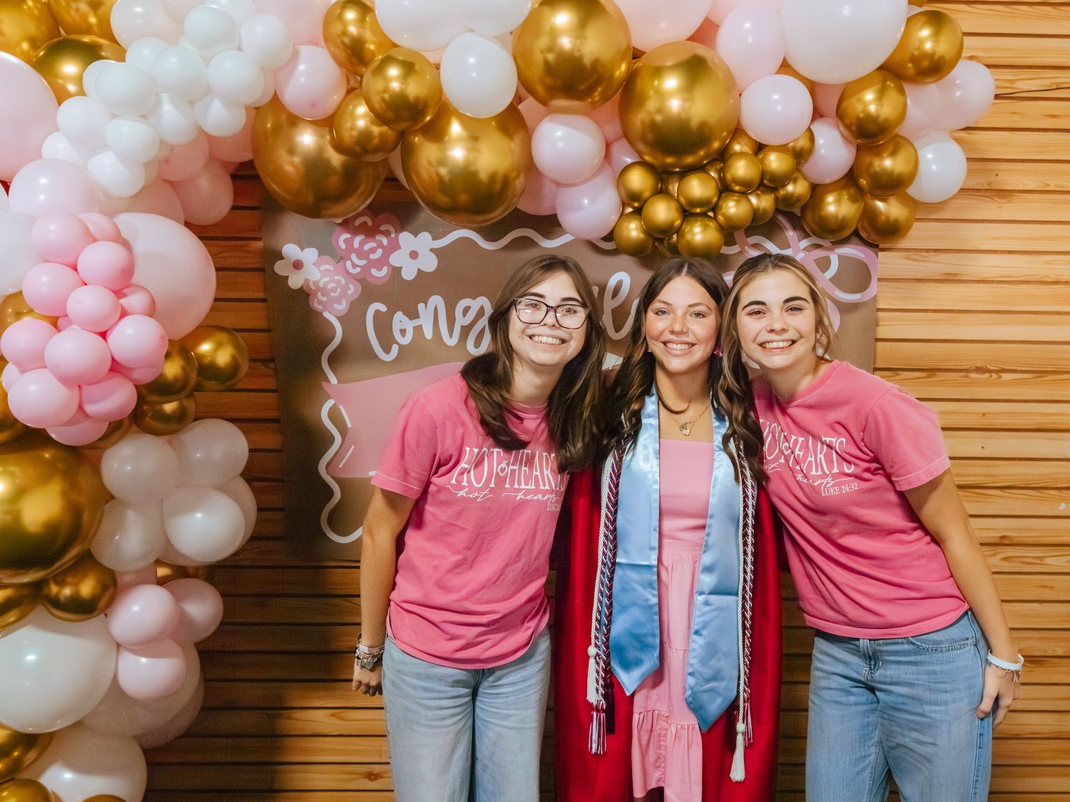 Three women pose at a graduation party with pink and gold balloon arch and Congrats sign backdrop.