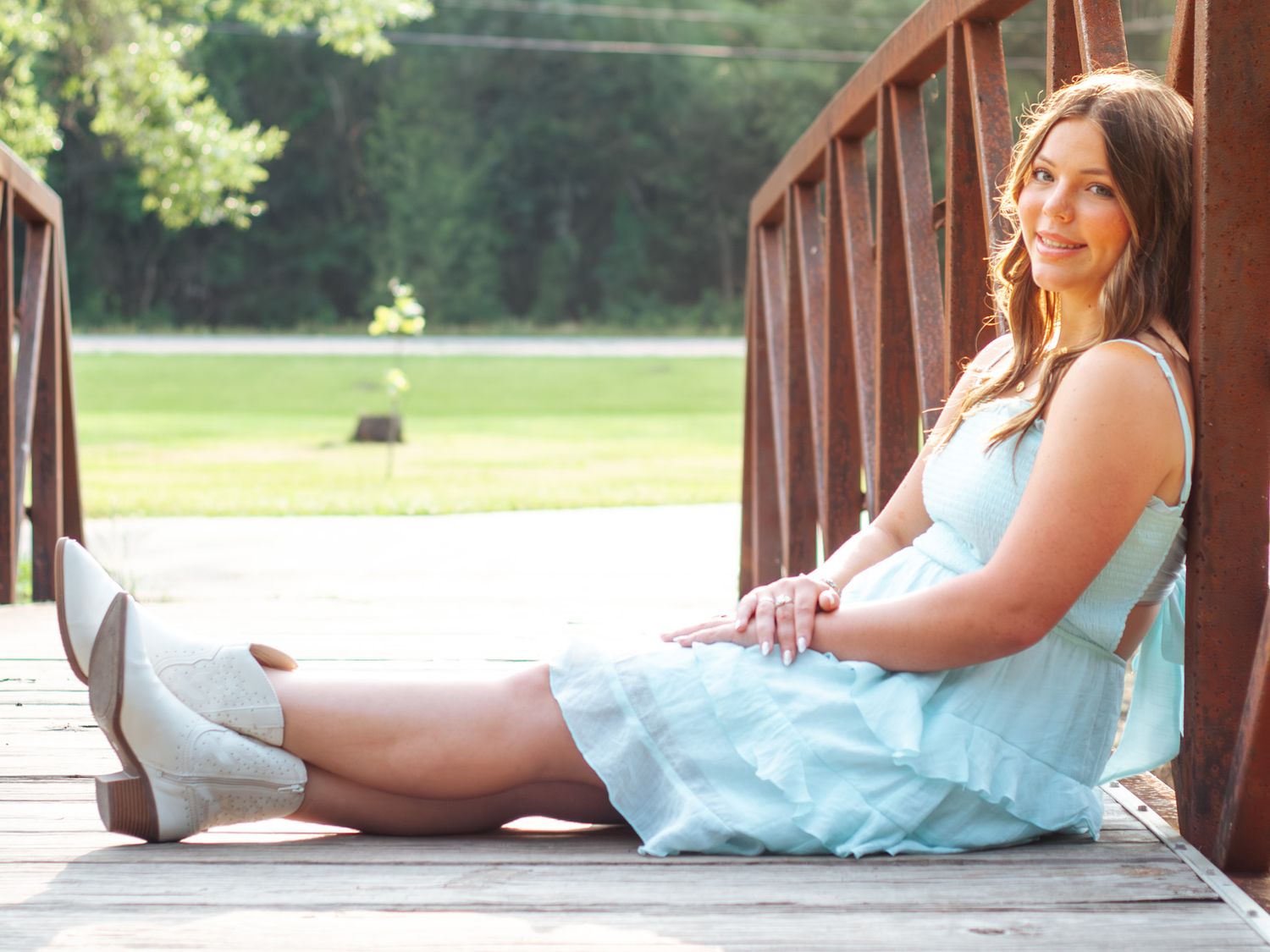 Young woman in light blue dress and boots sitting on wooden bridge outdoors.