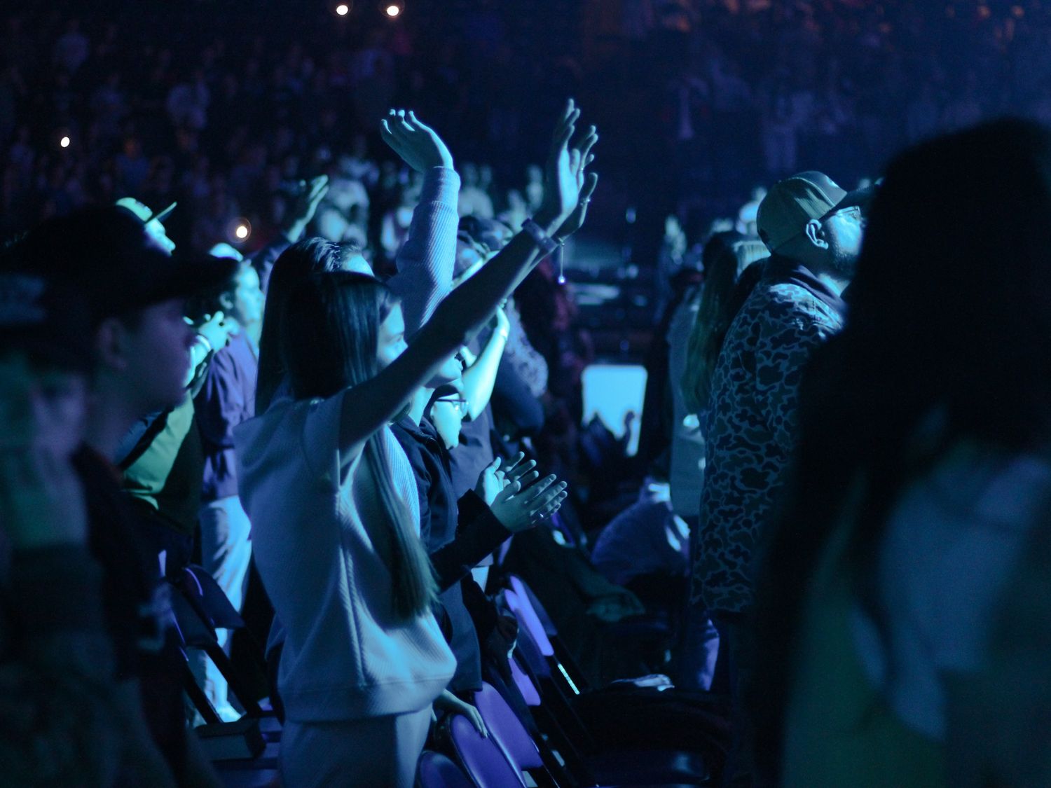 Enthusiastic audience members raising hands under blue stage lighting at a live concert event.