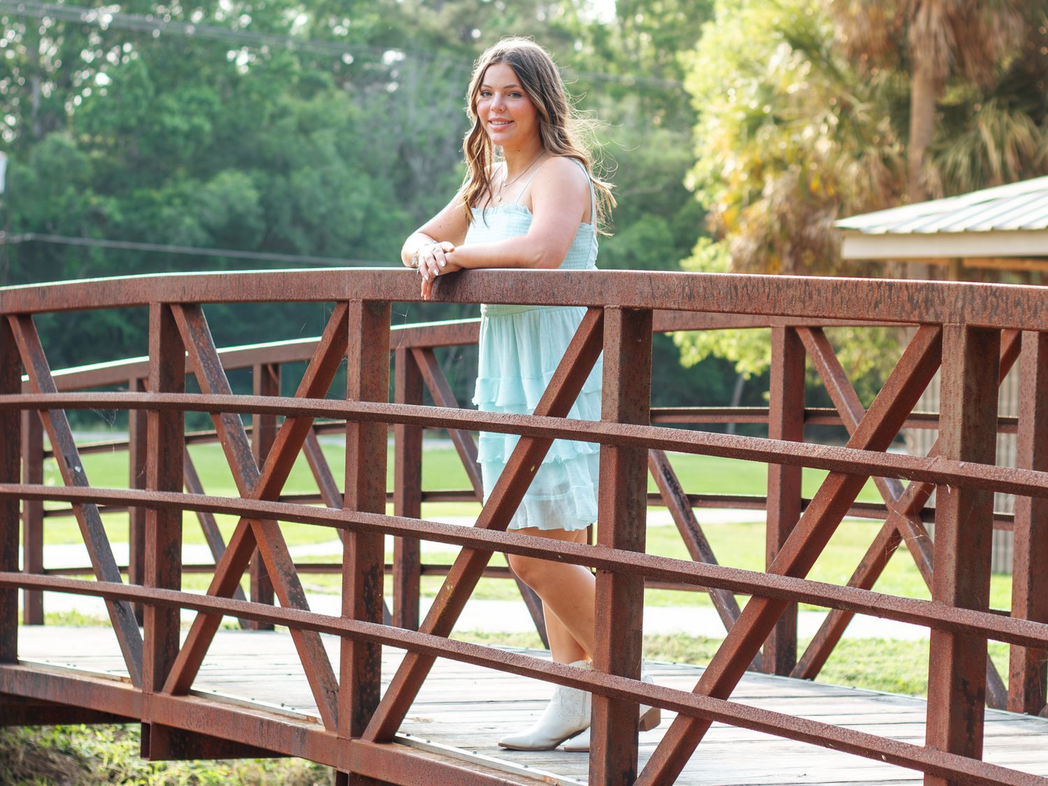 Young woman in light blue dress posing on a rustic wooden bridge surrounded by lush green trees.