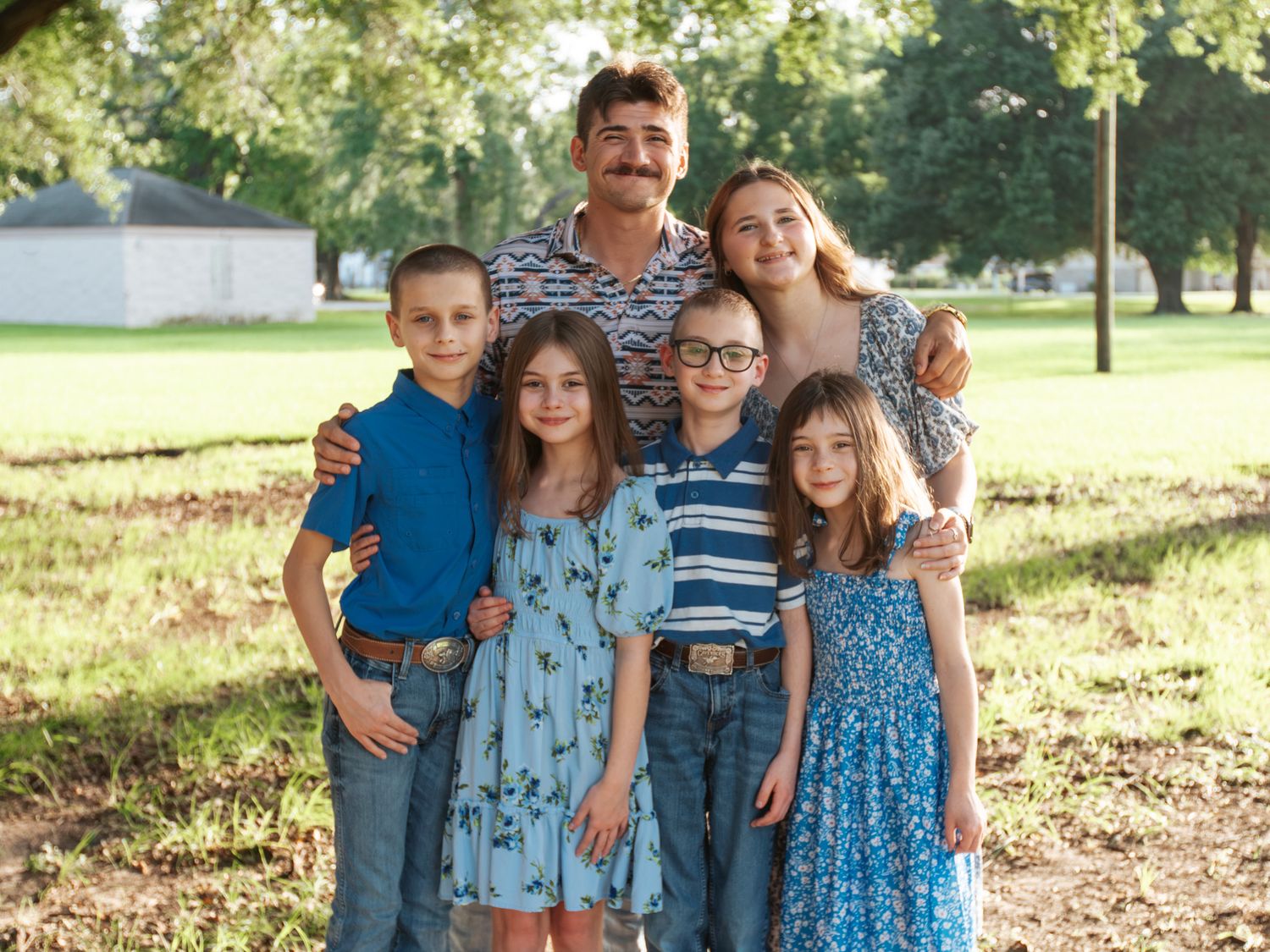 Family of six posing outdoors in a sunny park, parents with four children dressed in casual attire smiling together.