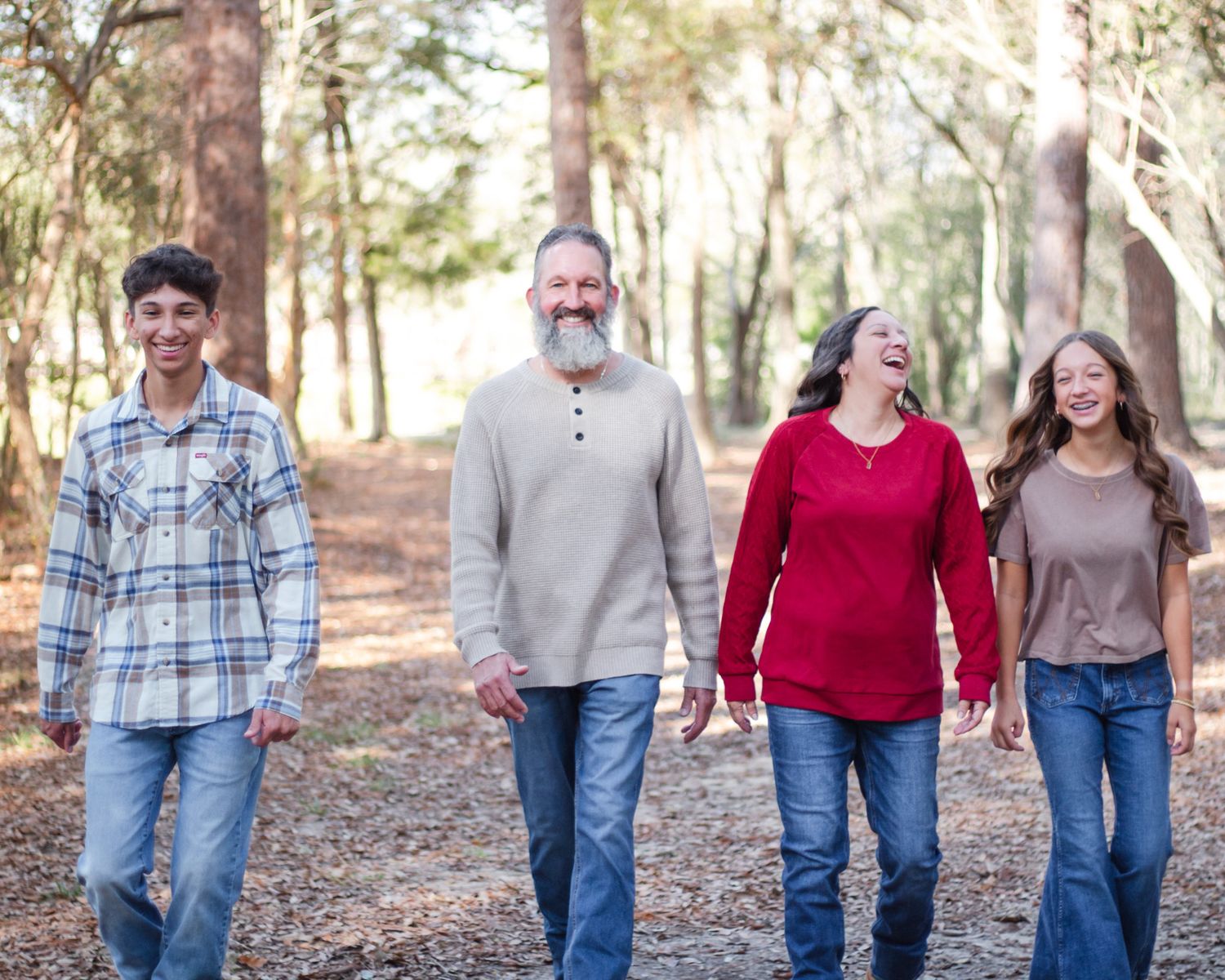 Family of four walking together on a forest trail, smiling and laughing among tall trees.