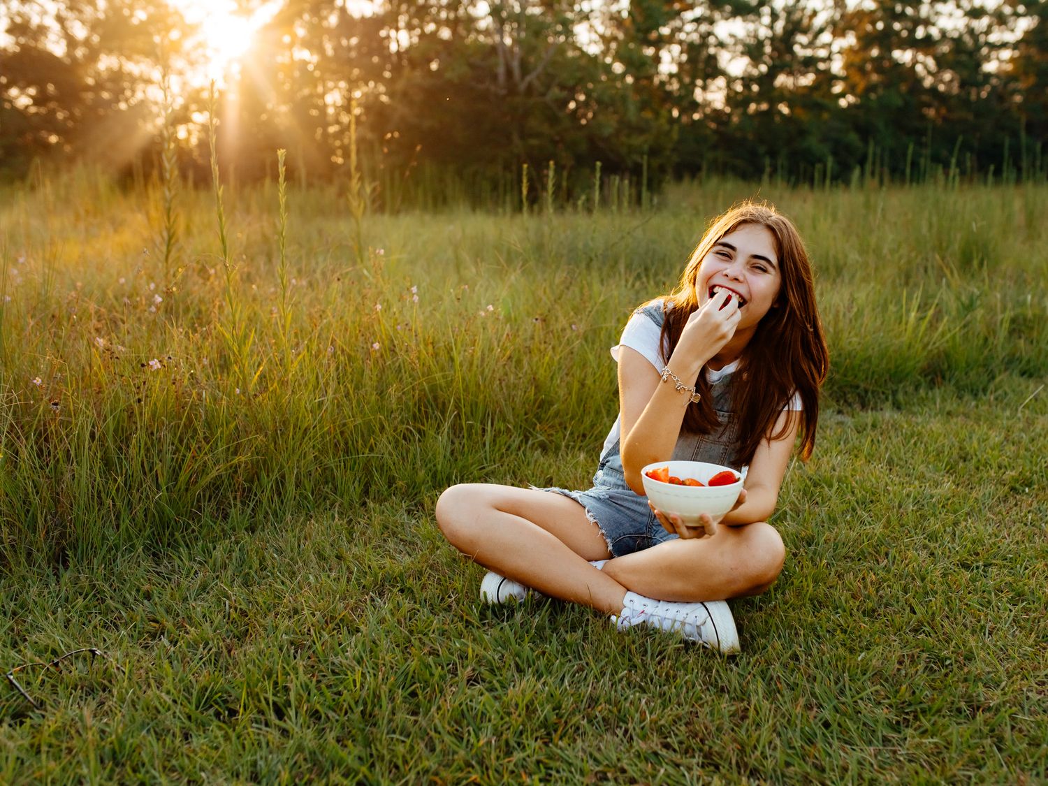 Young woman sitting cross-legged on grass eating strawberries from a bowl at sunset in a green field.