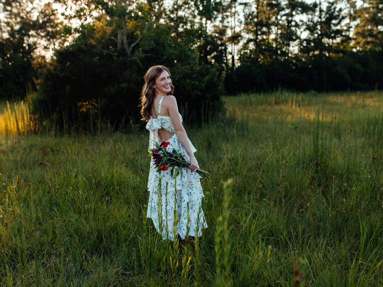 Bride in white lace dress holding bouquet stands in sunlit green meadow with forest backdrop at golden hour.