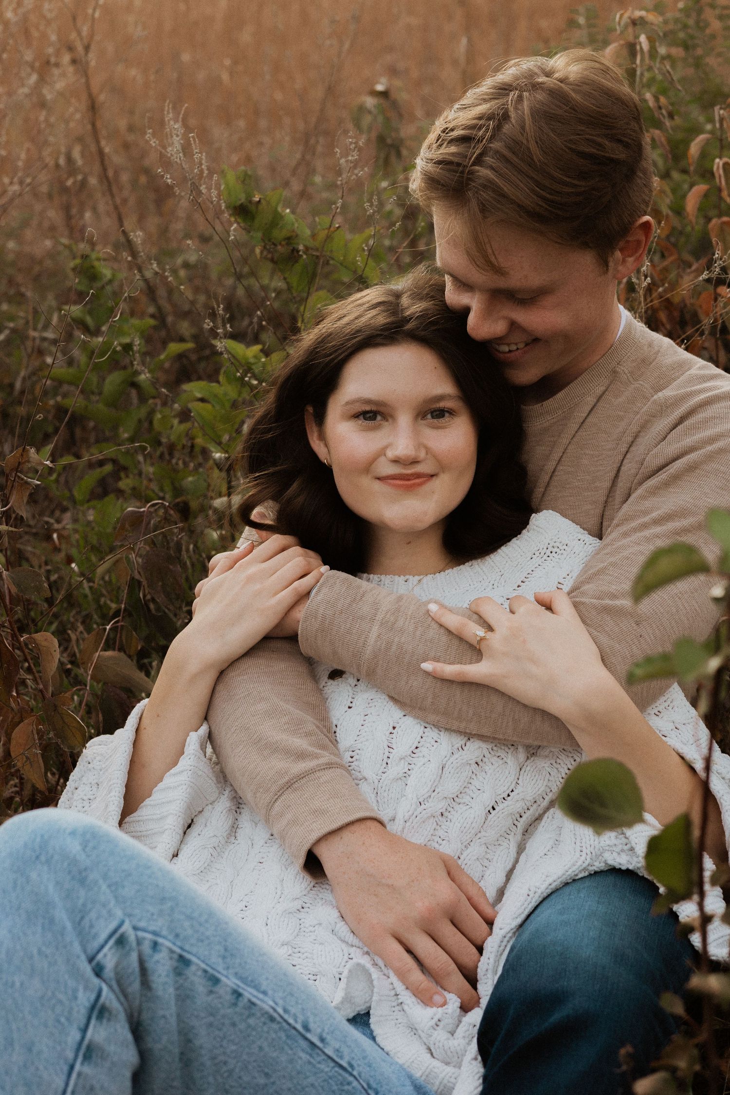 A couple relaxes together in casual attire among tall grass during an autumn photoshoot.