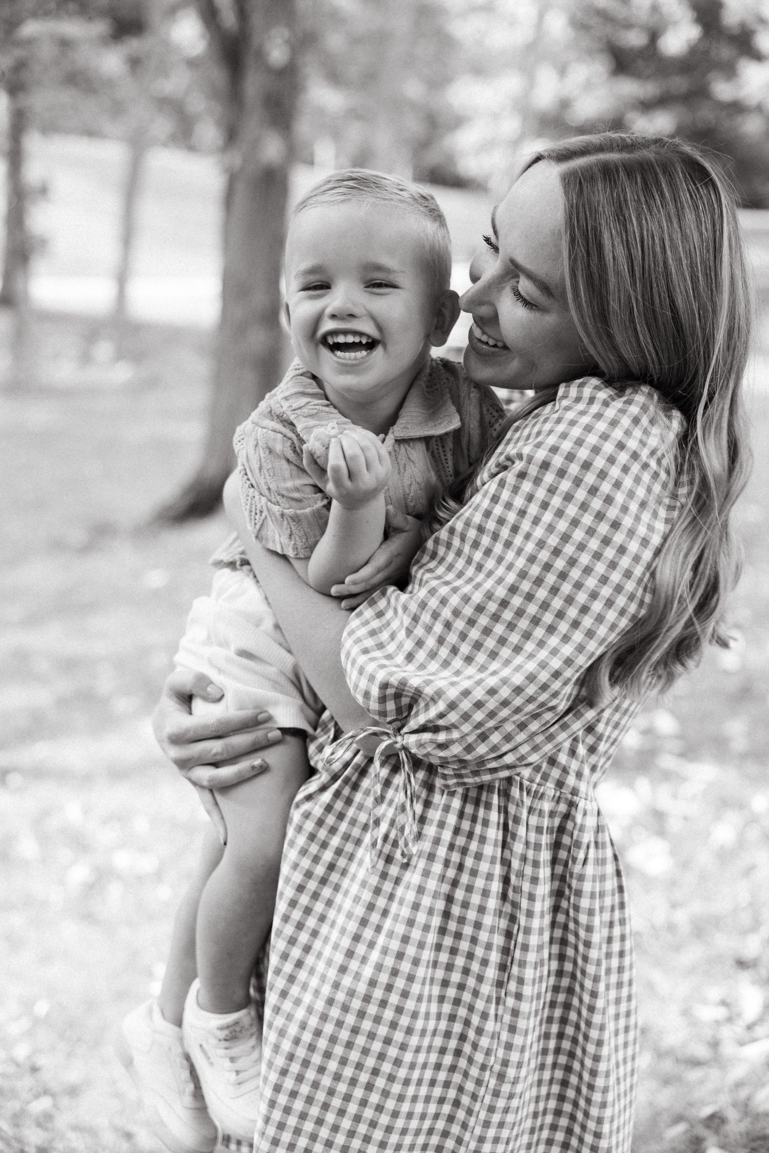 A joyful black and white portrait of a mother in a gingham dress holding her laughing toddler outdoors.