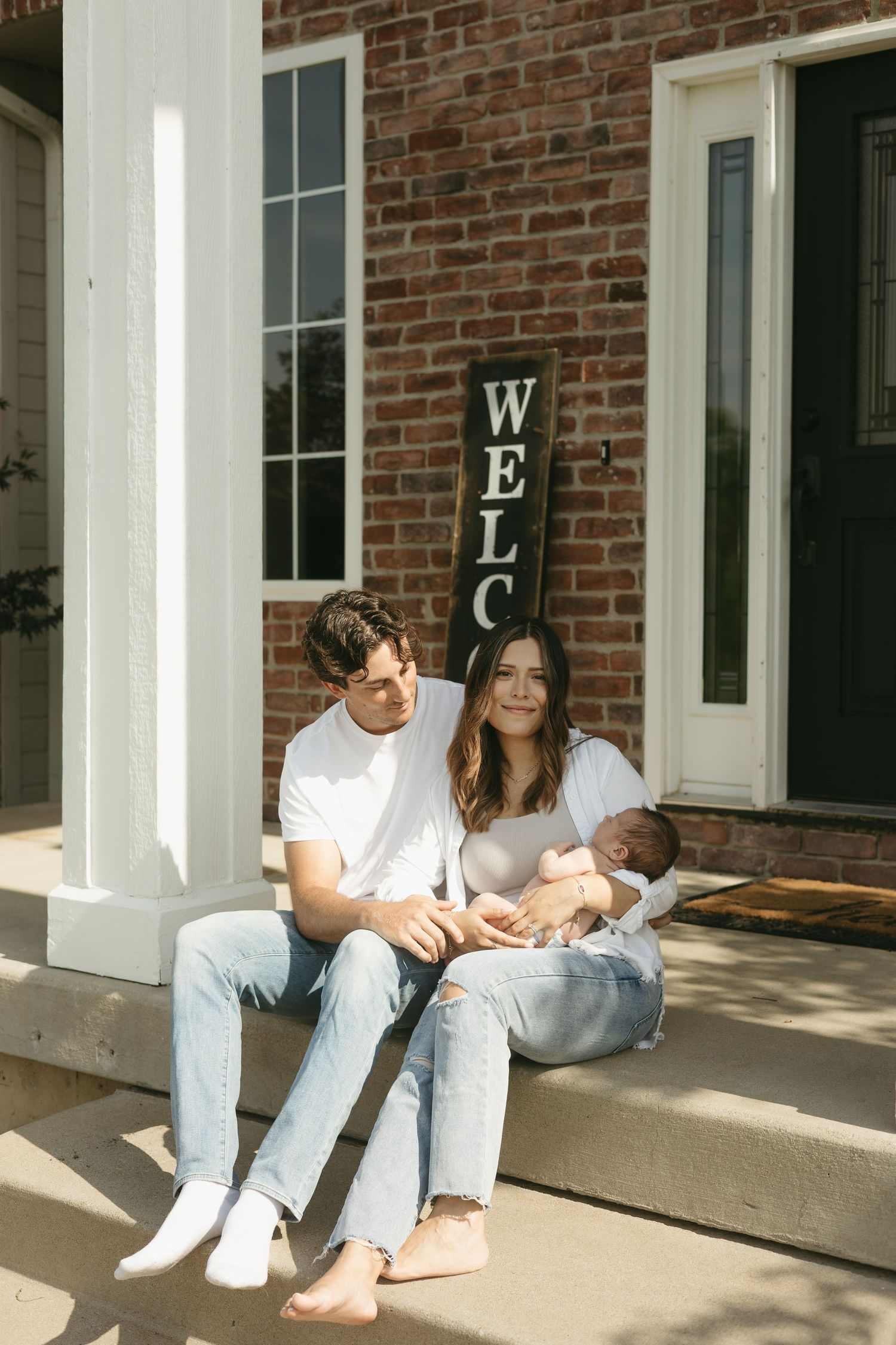 Couple in casual white outfits and jeans sitting on brick porch steps in sunlight.