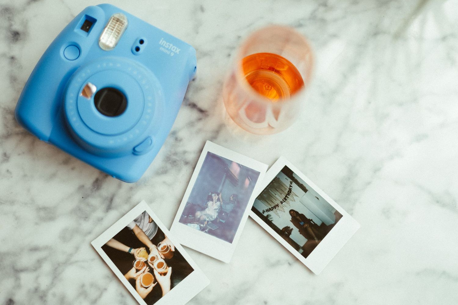 Light blue Instax camera with instant photos and orange film canister on marble surface.