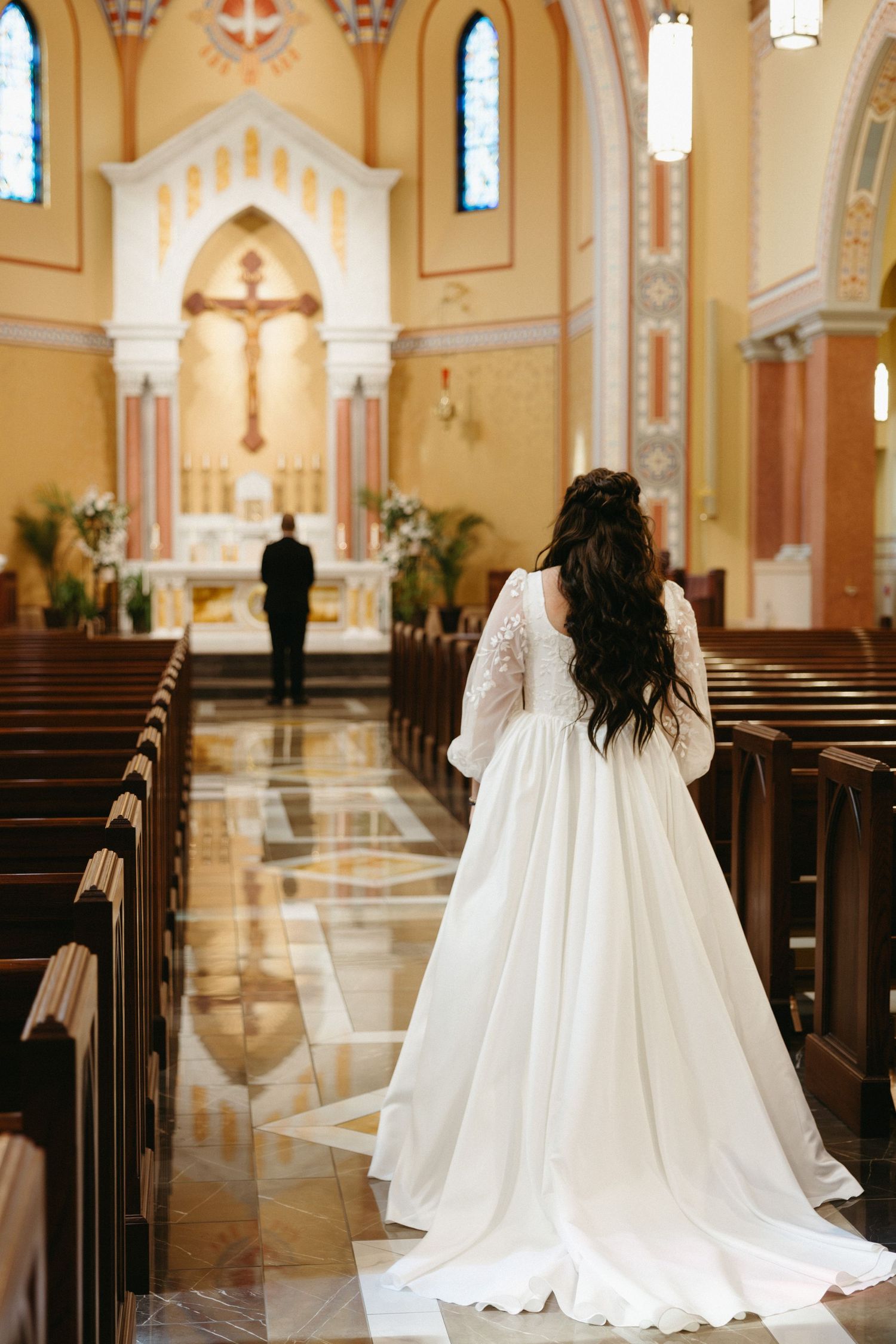 A flowing white wedding dress photographed from behind in a church aisle with ornate cream and gold architecture.